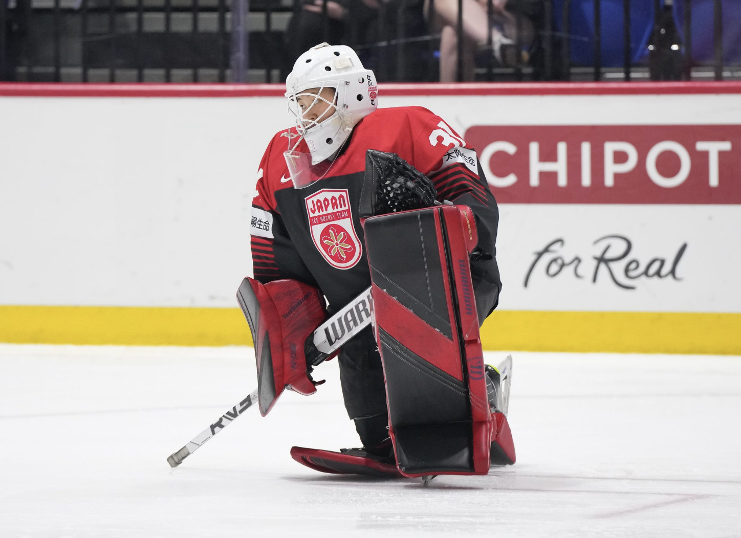 Riko Kawaguchi takes a knee in a faceoff circle and gazes up the ice. She is wearing a black and red jersey, white mask, and black and red gear.