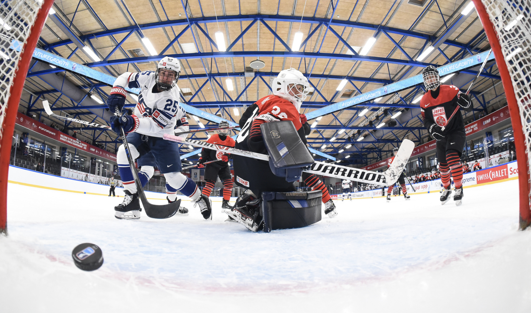 A view from inside the net as Carpenter scores on Masuhara. Masuhara is on her knees looking back and Carpenter is upright following through on her shot off to Masuhara's left. There are multiple Japanese skater in the background. Carpenter is in white, while the Japanese players are in black and red.