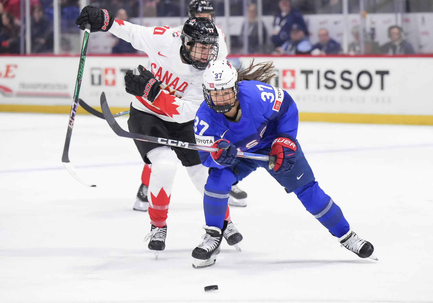 Murphy skates with the puck as Nurse chases. Murphy is more hunched over, while Nurse is standing a little more upright as she comes in from Murphy's right. Nurse looks like she is about to reach her stick around Murphy. Murphy is wearing blue, while Nurse is wearing white.