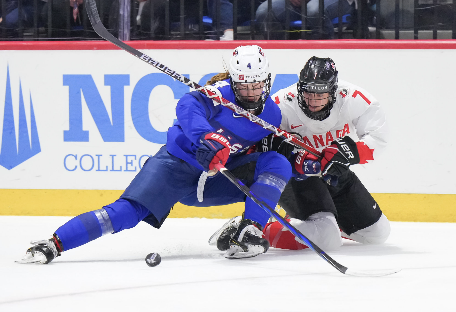 Harvey (left) and Stacey (right) battle over the puck. Stacey is on her kness, while Harvey has nearly fallen to hers as well. The puck sits in front of Harvey as Stacey reaches for it. Harvey is in blue, while Stacey is in white.