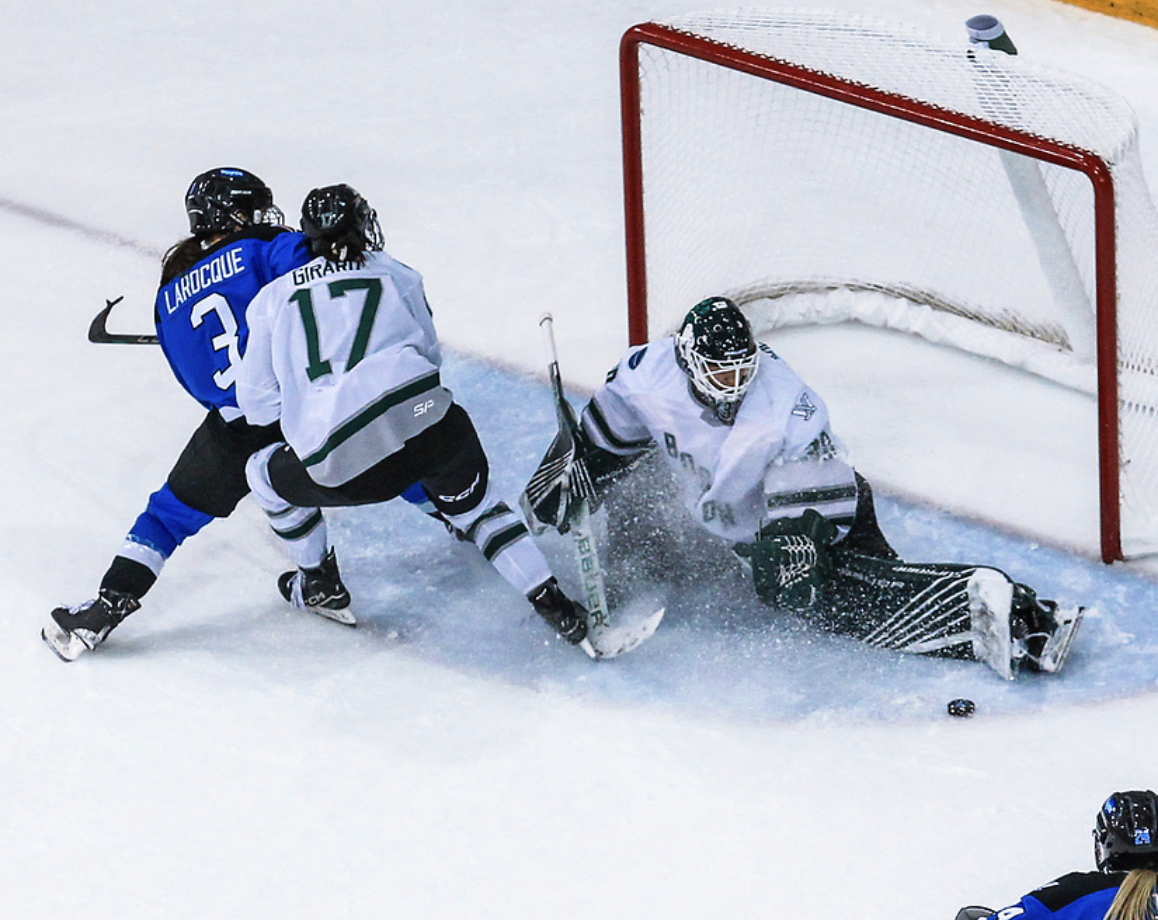 Girard and Larocque battle while Emma Söderberg makes a save. They're off to the left of the screen, with Girard maintaining contact with Larocque from behind. Söderberg is next to them on her knees, kicking her left leg out to make the save. The Boston players are in white, while Toronto is in blue.