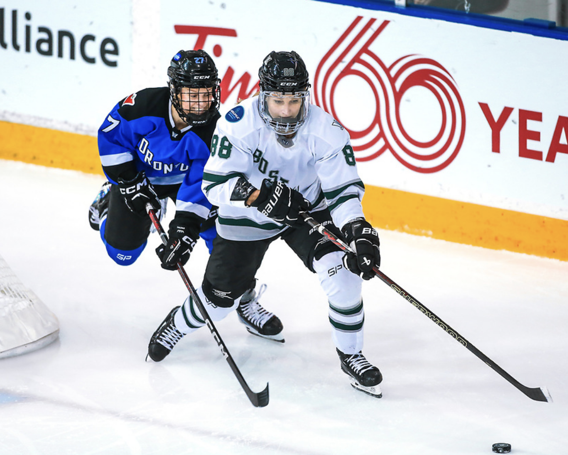 Maltais (left, back) tries to defend against Tapani (right, front). Both are slightly crouched while skating, and their sticks are in front of them. Tapani is carrying the puck. Tapani is in white, while Maltais is in blue.