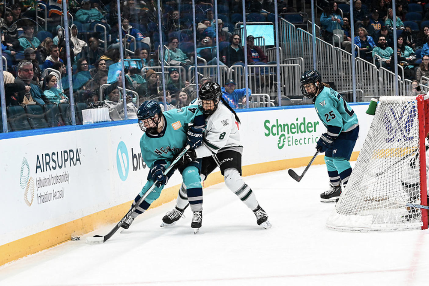 Adzija (right) battles for the puck with Baker (left) along the boards behind the New York net. Alex Carpenter looks on in the background. Adzija is in a white awa uniform, while Baker and Carpenter are in teal. 