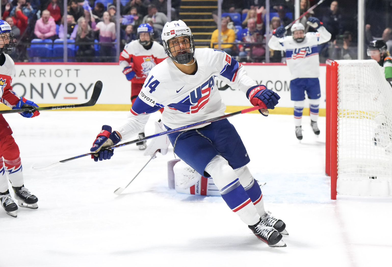 Laila Edwards skates away from the net while Kendall Coyne Schofield jumps in celebration in the background. They are both wearing white USA jerseys.