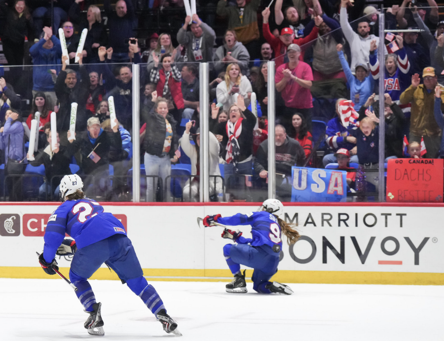 Simms celebrates fist pumps from one knee after scoring the overtime winner against Canada, while Taylor Heise skates to join her. They are both wearing blue USA jerseys.