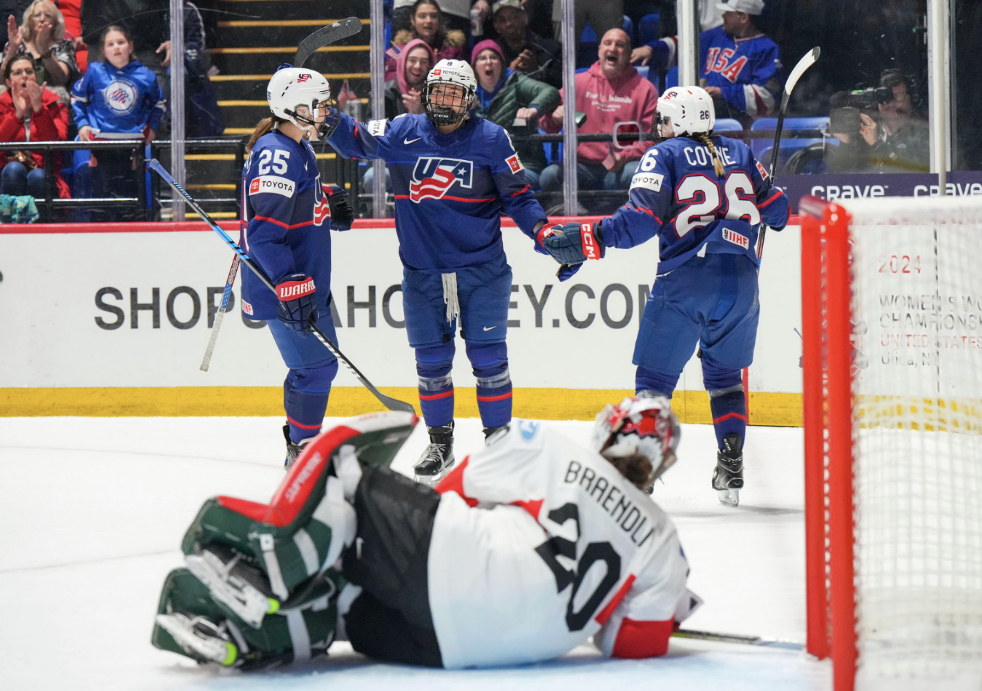 Winn celebrates her goal with a group hug featuring Carpenter and Coyne Schofield. They are wearing blue USA jerseys.