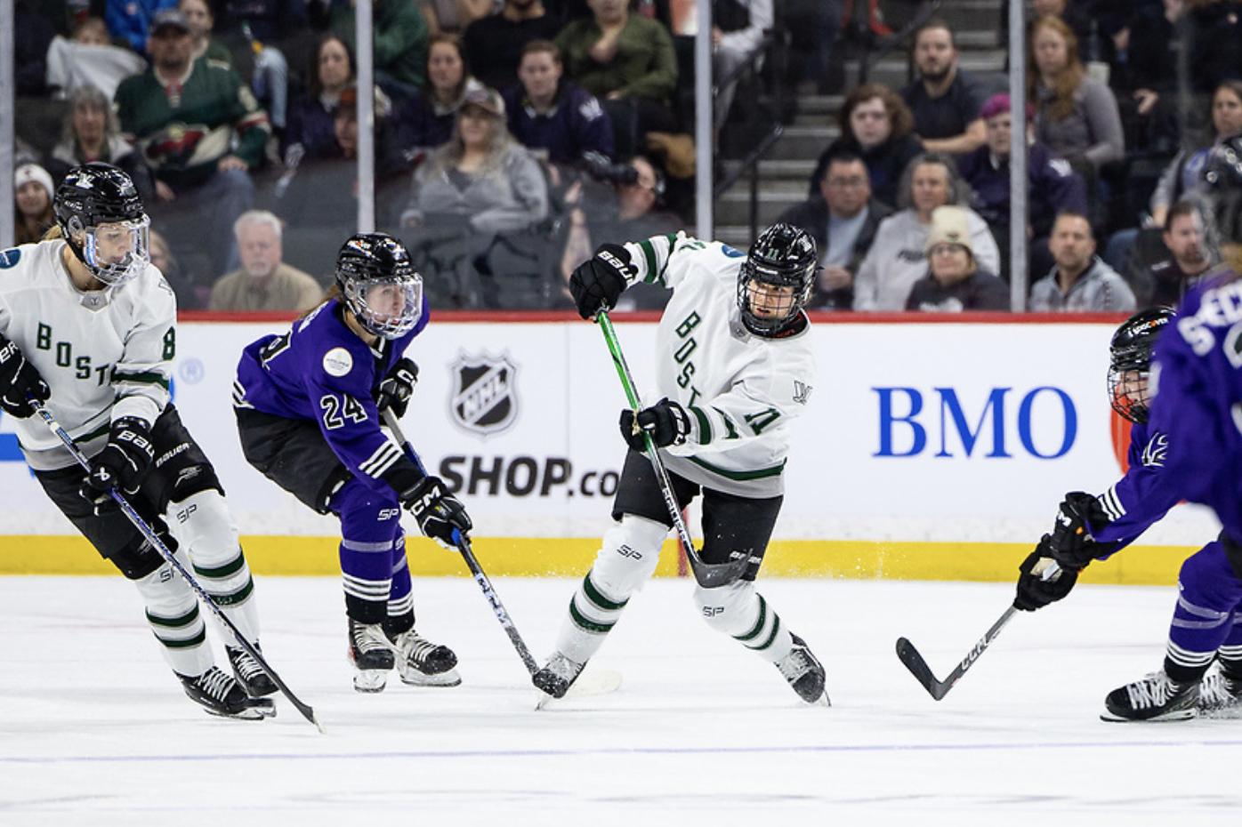 Alina Müller drops her shoulder and raises her stick as she follows through on a pass to a teammate. She is wearing a white away jersey, and there are several Minnesota players near her.