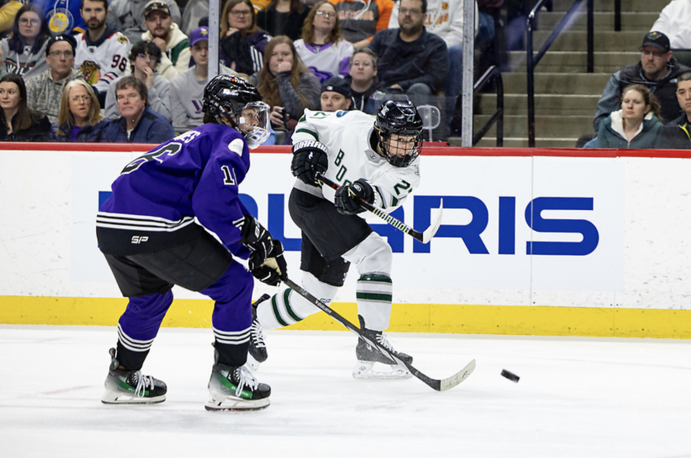 Darkangelo follows through on her shot on goal as Jaques tries to defend. Jaques is turning to skate forward while Darkangelo is mid-stride. Darkangelo is in white, while Jaques is in purple. 