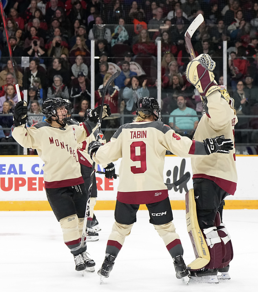 Poulin (left, arms raised), Tabin (center, arms to the side), and Desbiens (right, arms raised) celebrate a win with a group hug. They are in cream away uniforms.