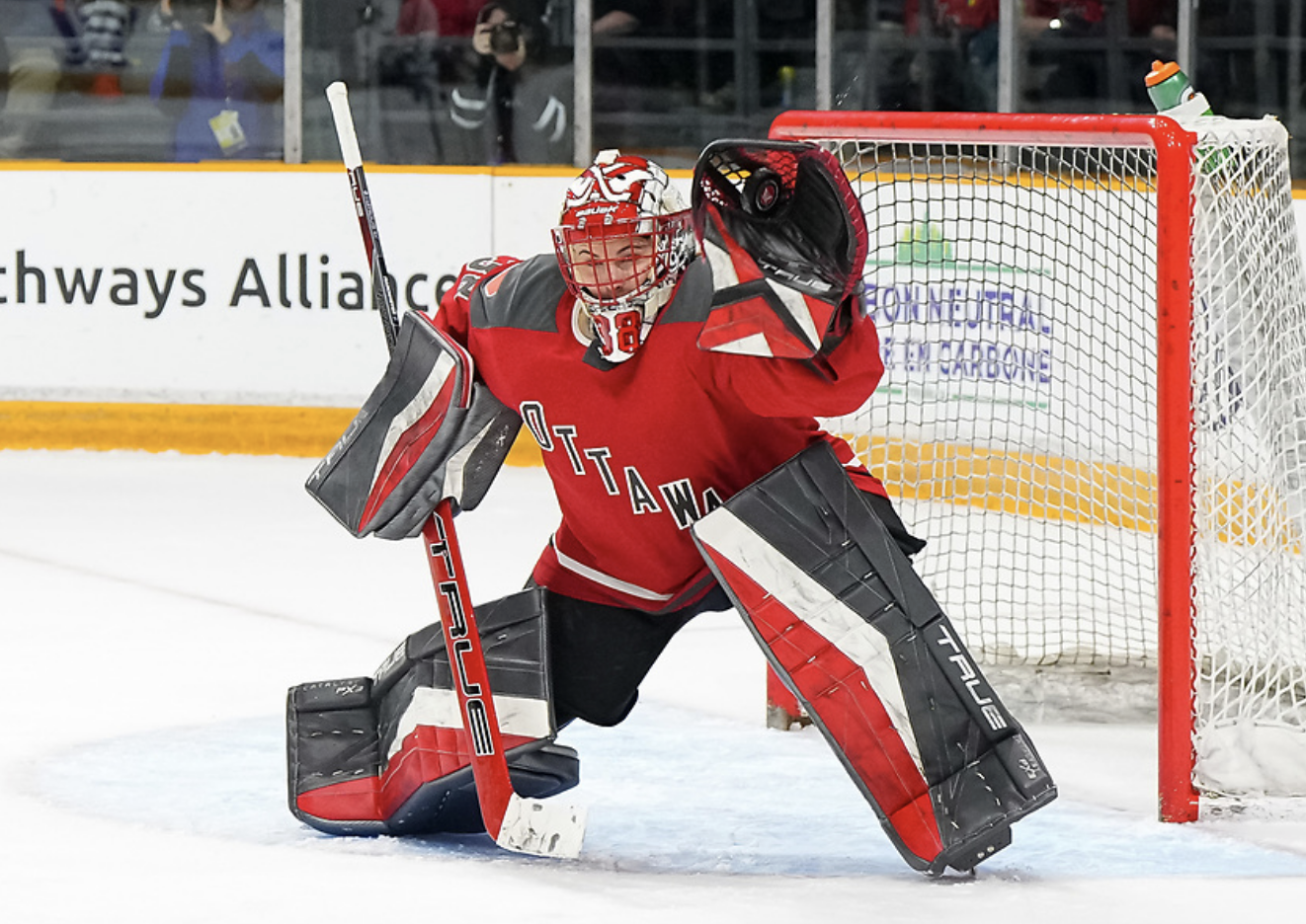 Maschmeyer makes a glove save against Boston. Her glove is raised as the puck enters it, while her stick is down and five hole wide open since she is lunging up to the left. She is in red and black goalie gear and a red home uniform.