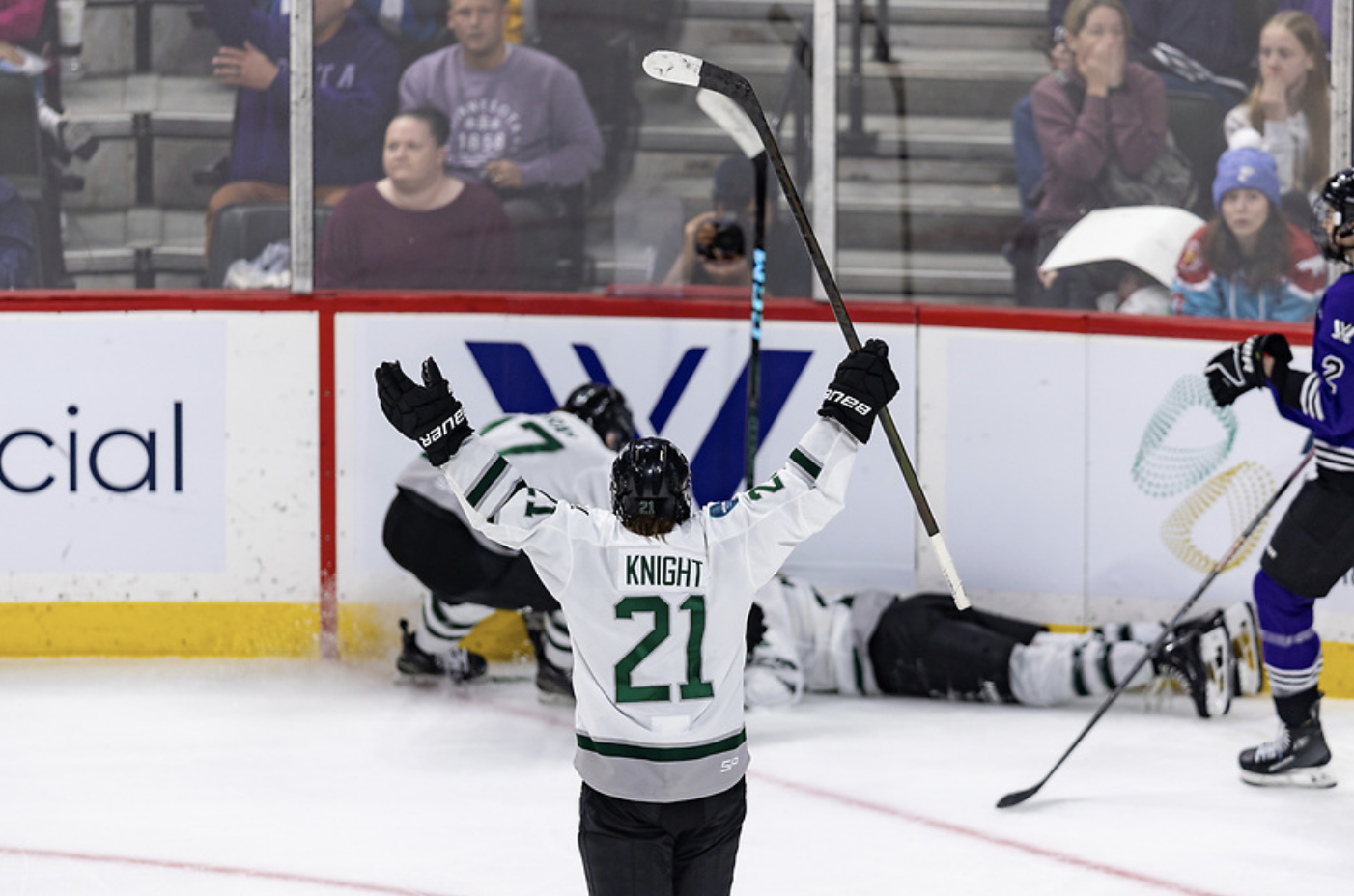 Knight raised her arms to celebrate Brandt's buzzer-beater goal. Brandt is laying on the ice in the background. They are wearing white away uniforms.