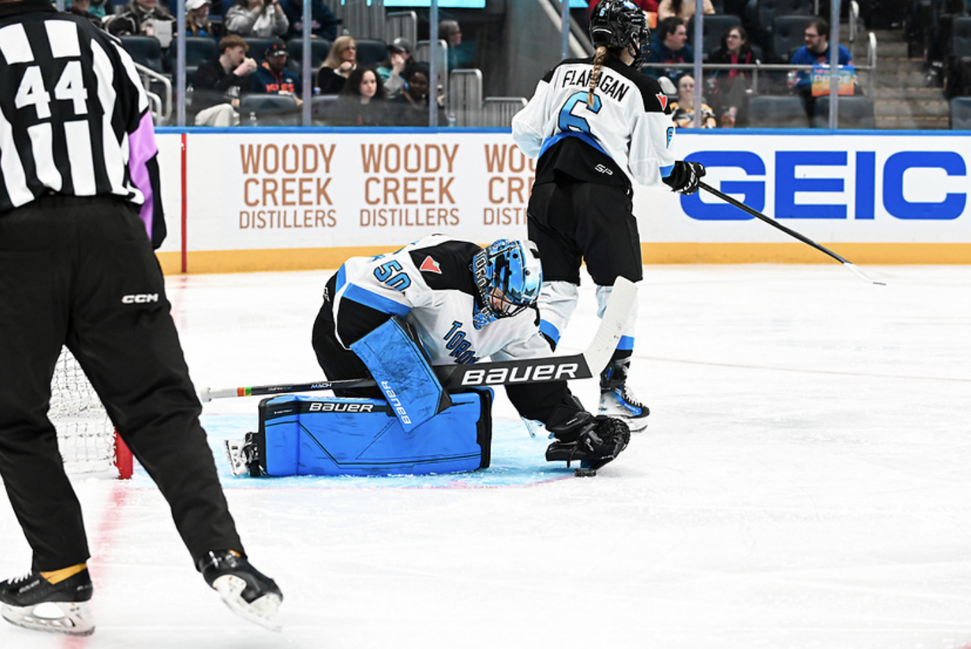 Campbell snaps up a puck during a game against New York. She is hunched over on her knees, and picking up the puck from the ice. She is in her blue mask and goalie pads, plus a white away uniform.