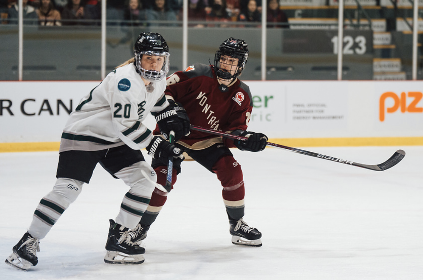 Brandt (left) and Bujold (right) battle for positioning at the top of the crease. Both are watching a play to their right and have their sticks at their waists. Brandt is in white, Bujlod in maroon.