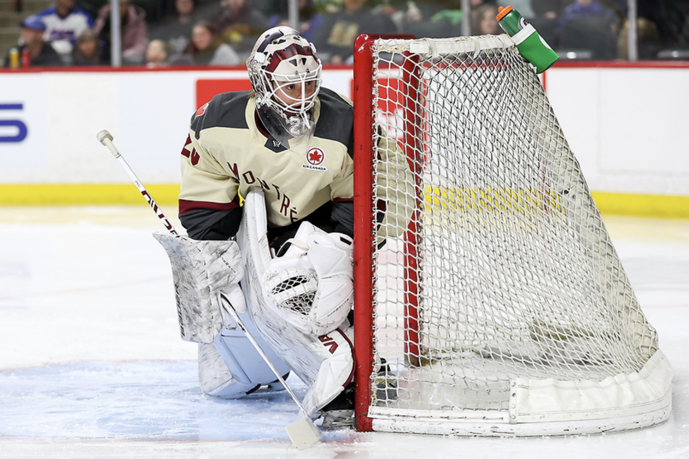 Chuli crouches while tracking the puck behind the net. She is wearing a cream away iniform, white pads, and a maroon and off-white mask.