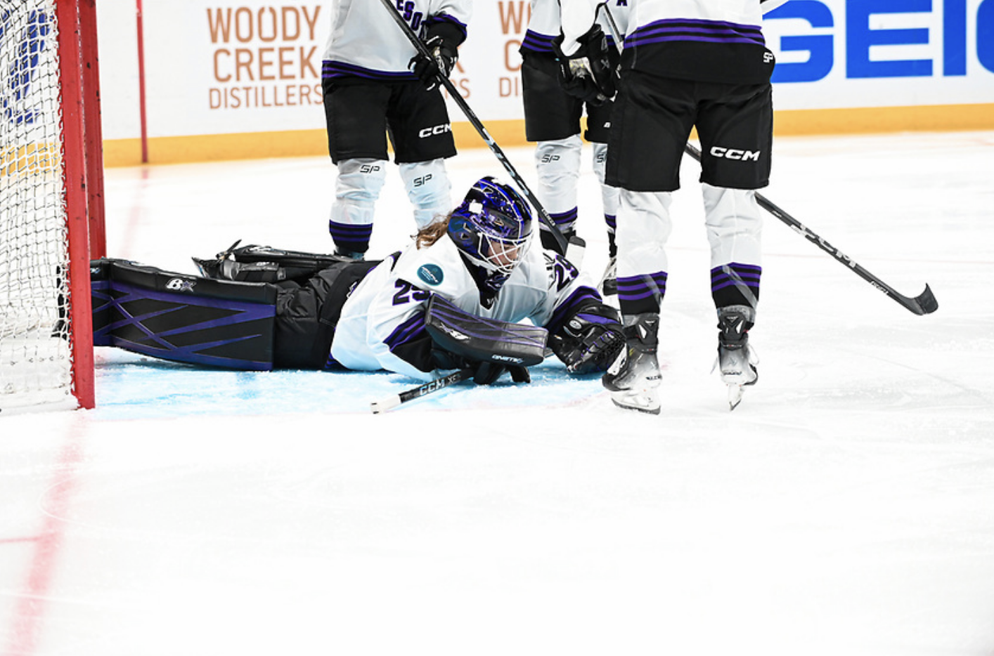 Hensley lays almost flat on her stomach in her crease after making a save. Three of her teammates stand around her, only visible from the waist down. They are all wearing white away uniforms.