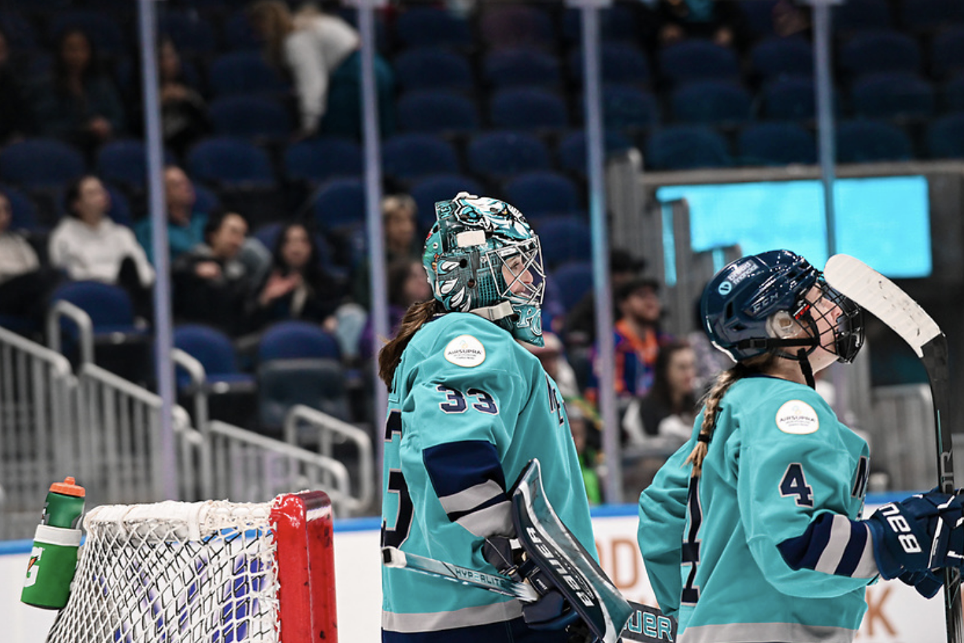 Post (left, in front of the net) and Baker (right, in front of Post) look up ice. They are wearing teal home jerseys.