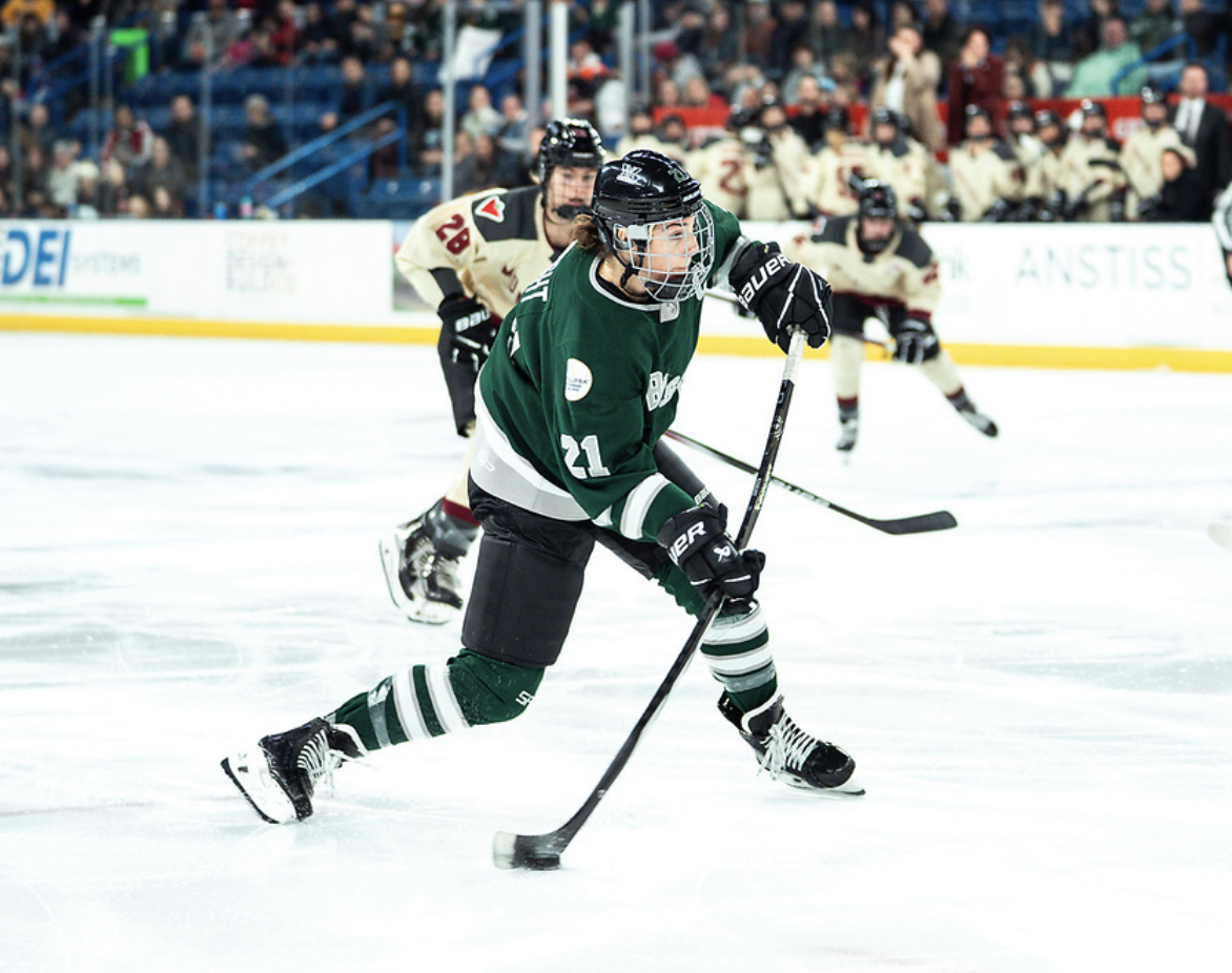 Hilary Knight's stick is heavily flexed as she loads up a shot. She is in the middle of shifting forward (onto her left leg). She is wearing a green home uniform.