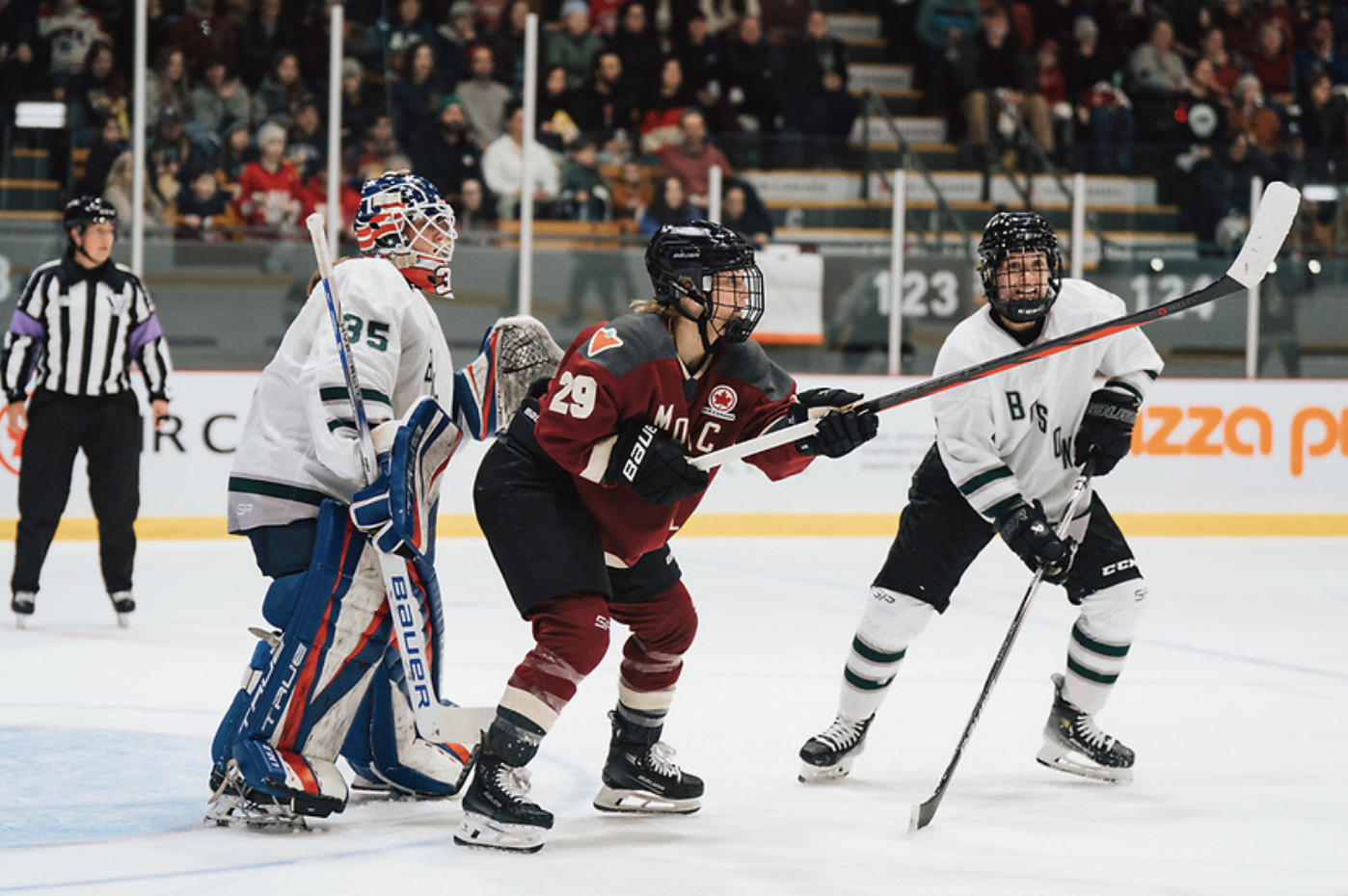 Poulin crouches and raises her stick to knock the puck out of the air in front of Frankel. Rattray looks on preparing to defend. Poulin is in maroon, while Frankel and Rattray are in white. 