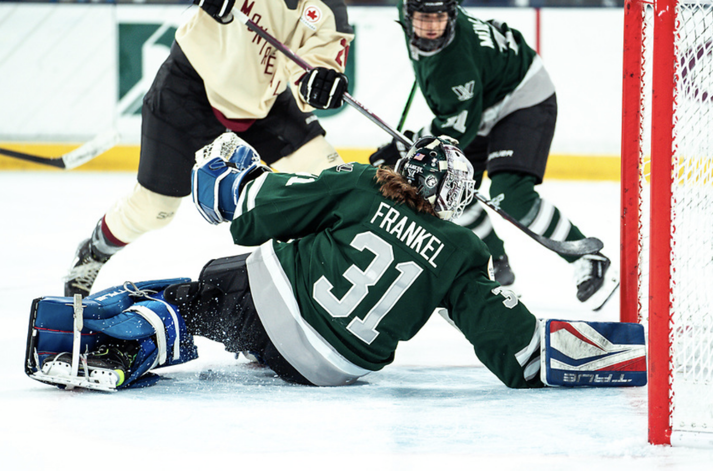 A from-behind shot of Frankel diving back towards her net to make a save with her blocker on the ice. She is wearing a green home uniform, green/white mask, and her USA pads.