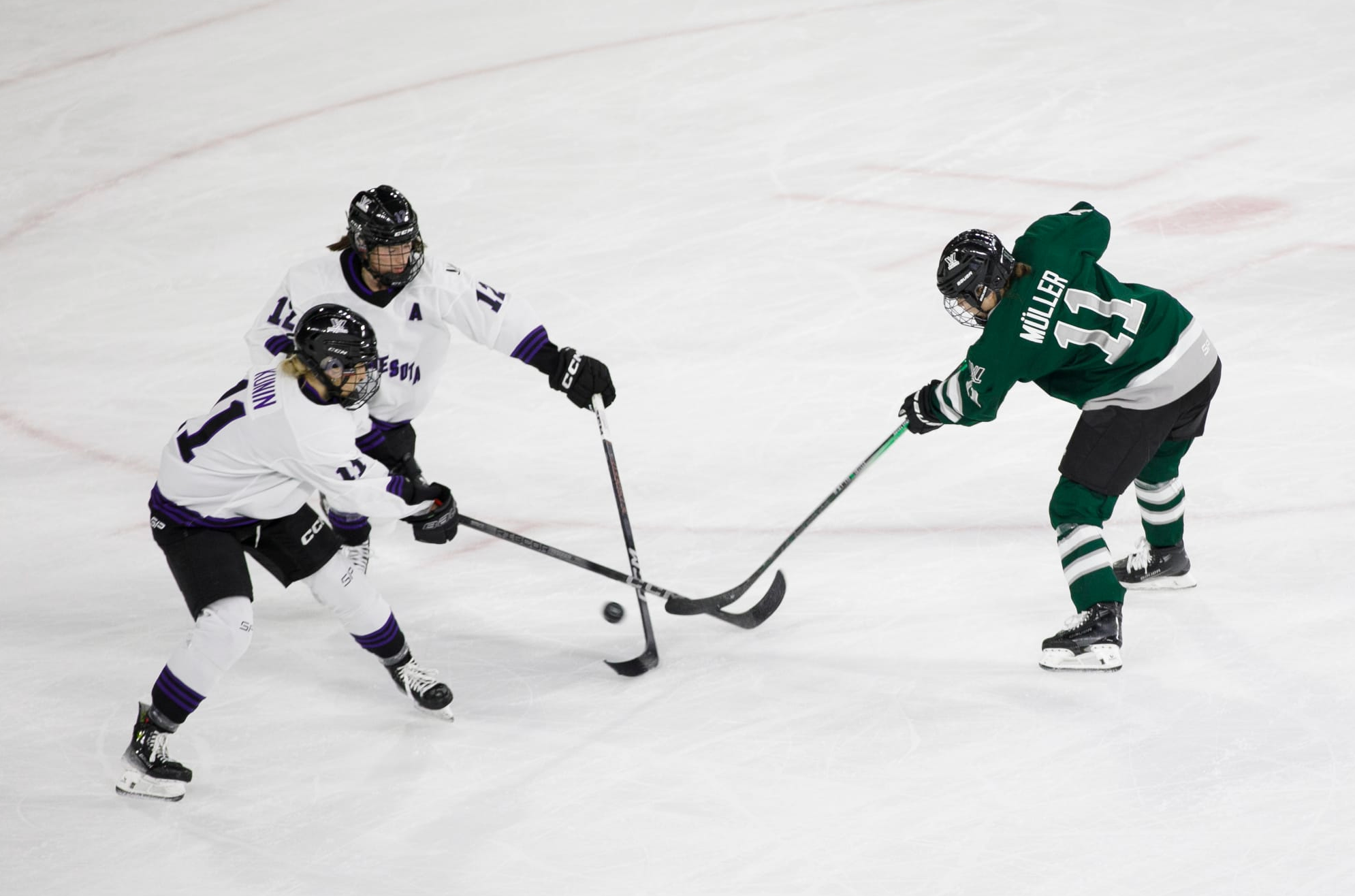 Müller (right in green) takes a shot through two Minnesota defenders (left in white). They're all leaning forward with sticks outstretched towards the puck.