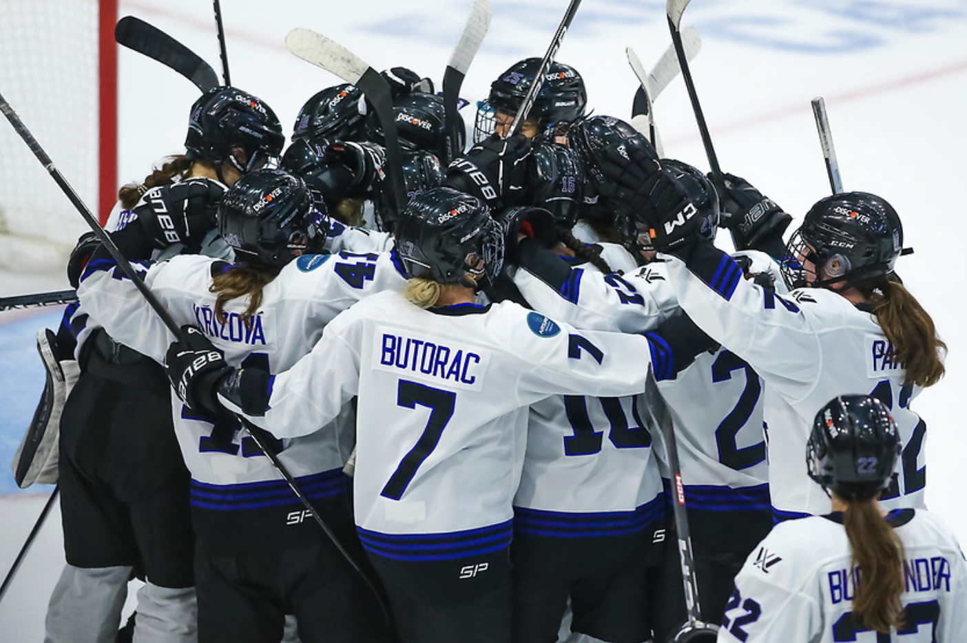 All Minnesota players celebrate with a big group hug in front of Maddie Rooney's net. They're wearing white away uniforms.