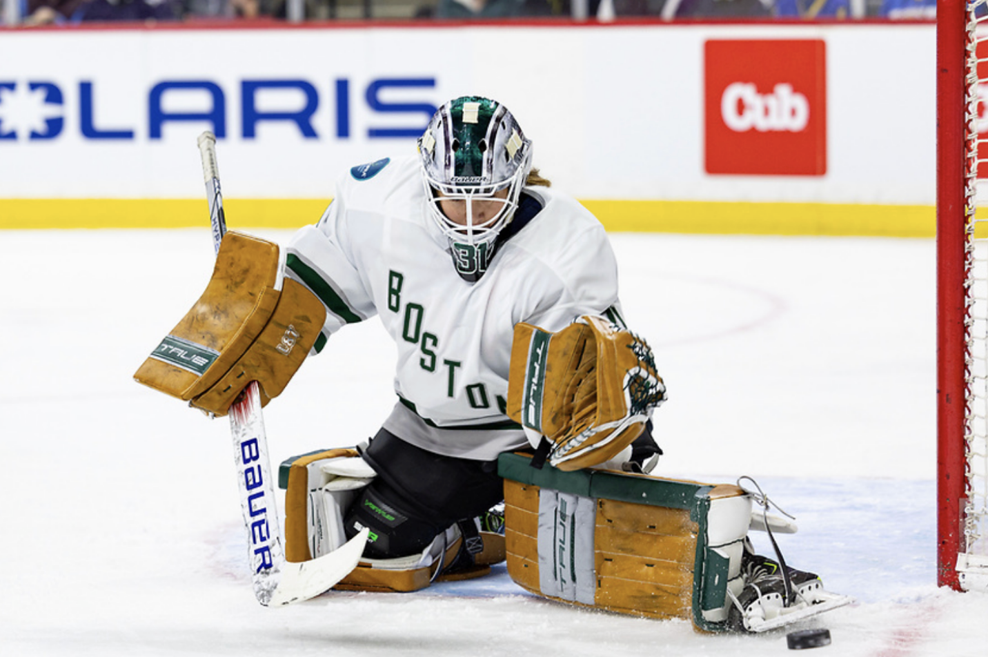 Frankel kicks the puck out to her left while making a save from the butterfly position. She is wearing a white jersey along with her green/white mask and tan pads.