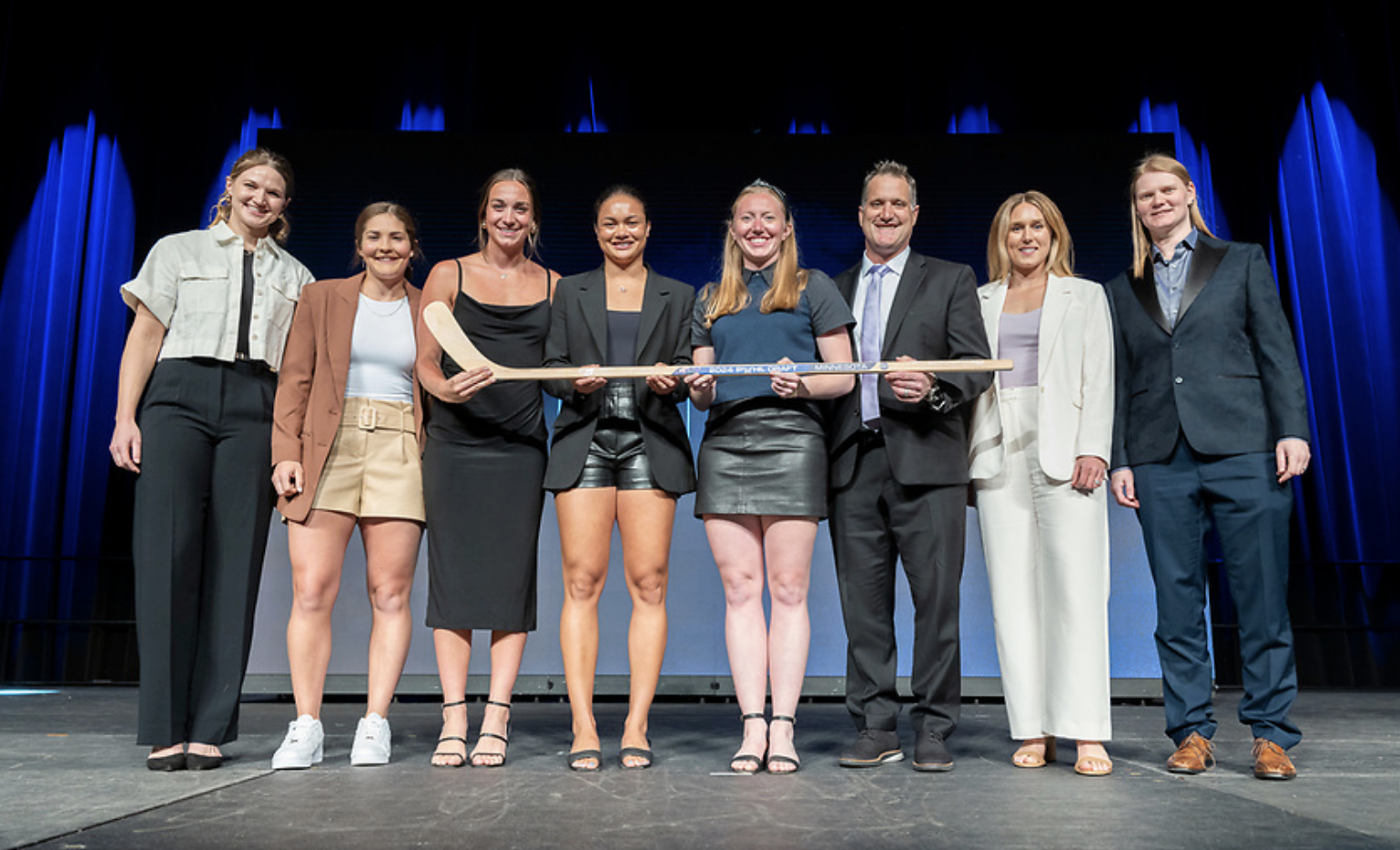 Eight representatives of PWHL Minnesota pose for a photo on stage while holding a wooden stick at the 2024 draft.