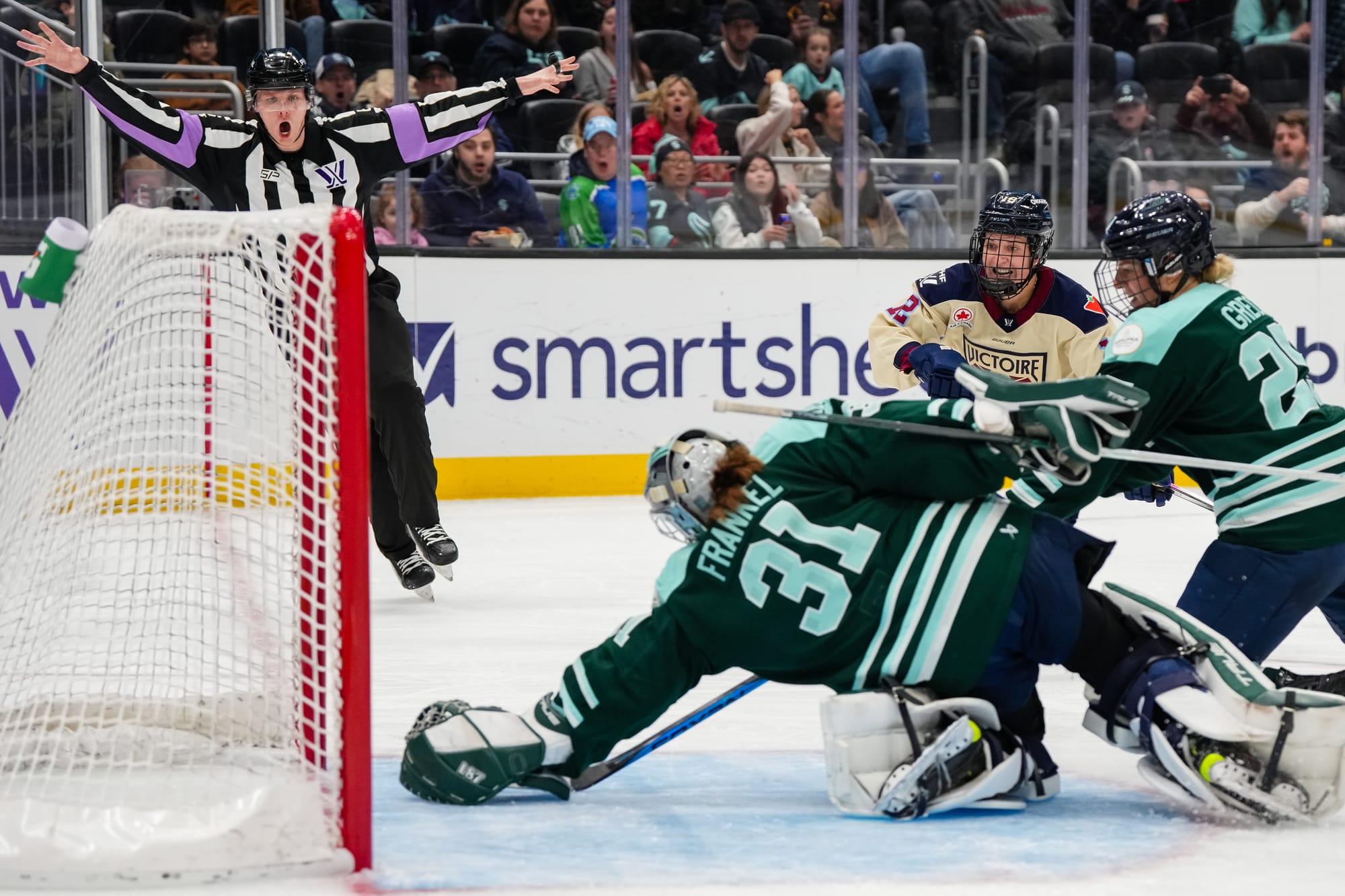 Frankel dives back with her arm outstretched. Emma Greco is also lunging back hoping to get a stick on the puck. A Montréal player looks on excitedly, but the referee on the goal line is signaling no goal. The Fleet players are in green, while the Montréal player is in a cream away uniform.