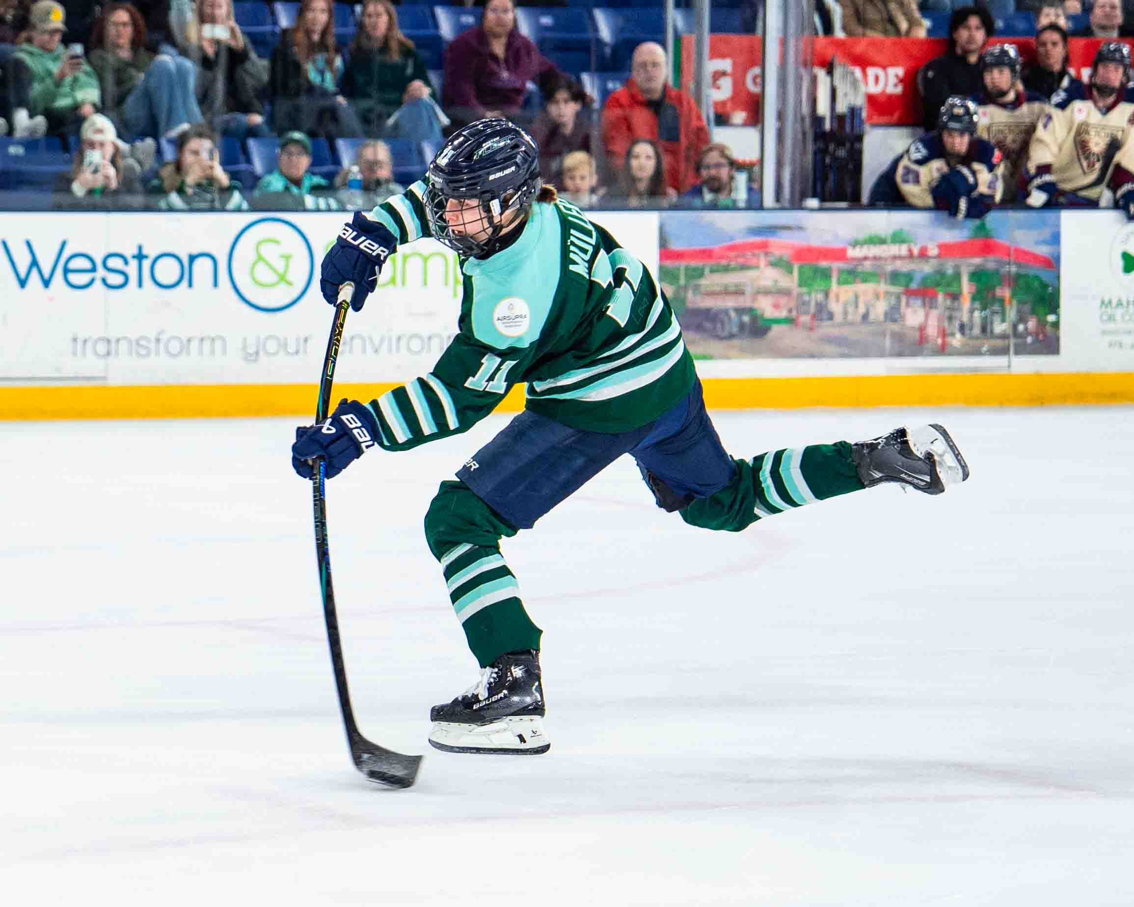 Müller leans forward, flexing her stick as she prepares to release a shot. She is wearing a green home uniform.