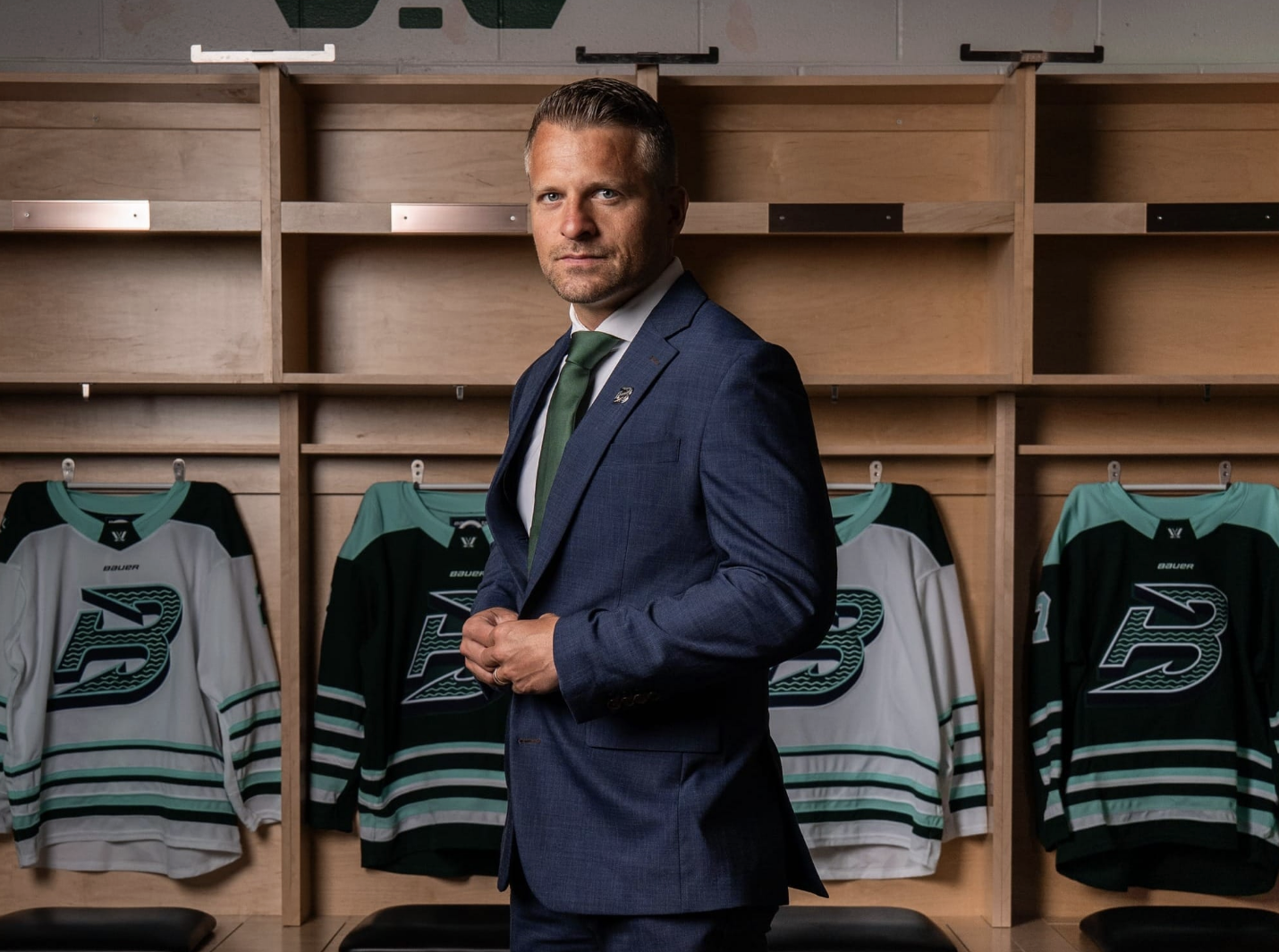 Sparre gazes into the camera while standing in front of home and away Fleet jerseys hanging in locker stalls. He is wearing a navy blue suit.
