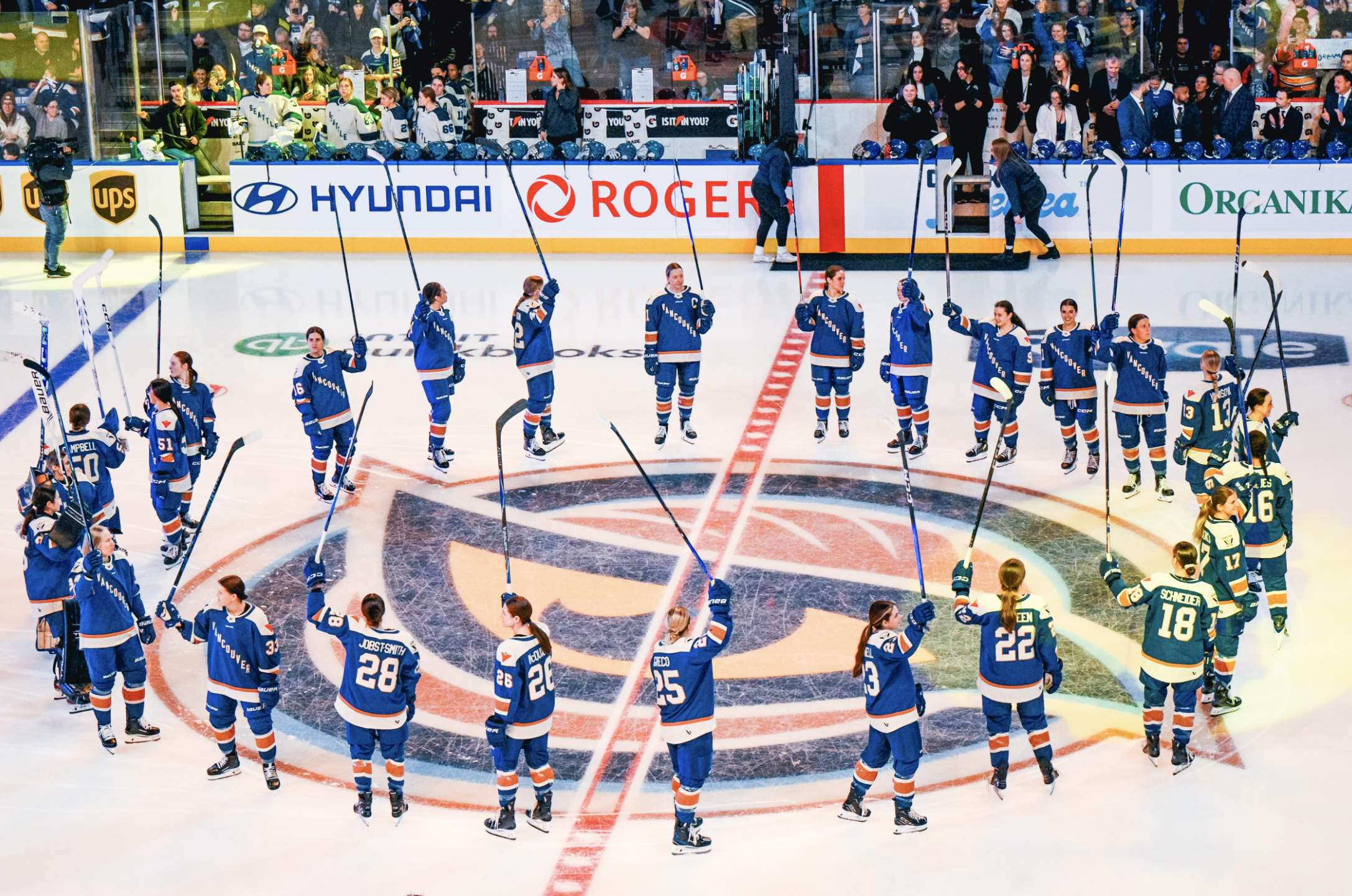 The Goldeneye stand around the center ice circle, which features their logo, raising their sticks in salute of the crowd. They are wearing their blue home uniforms.
