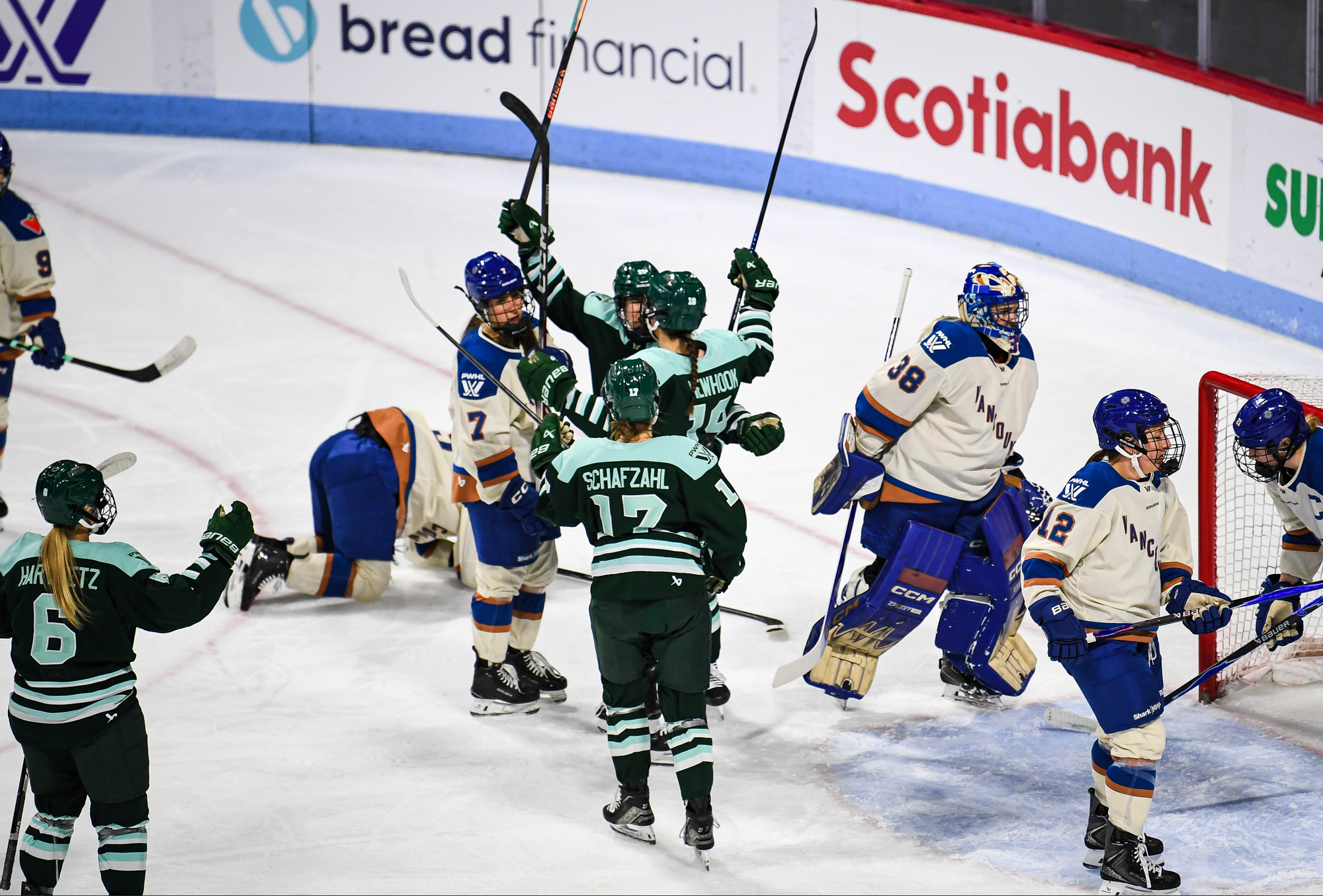 Newhook raises an arm in celebration right outside the Vancouver crease as her teammates gather around her for a group hug. They are wearing green home uniforms, while the Goldeneyes players in frame are wearing cream away ones.