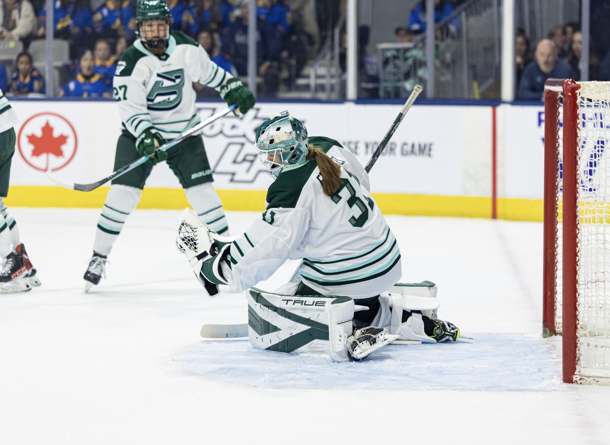 On her knees in her crease, Frankel leans forward as she clutches the puck in her glove. She is wearing a white away uniform.