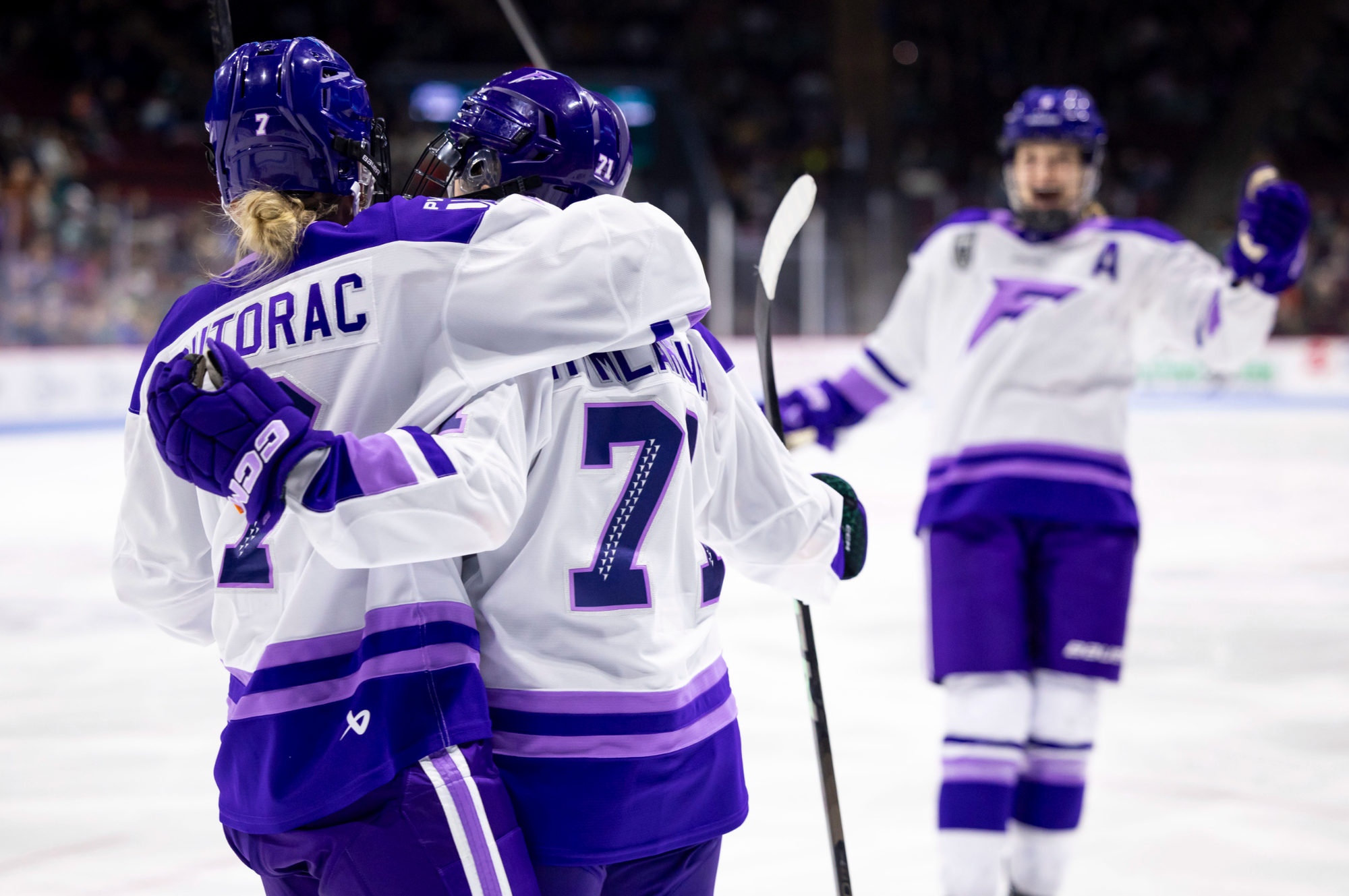 Three Minnesota players celebrate their goal with a group hug. Two have their backs turned to the camera, while the other is facing the camera but out of foucs. They are wearing white away uniforms.