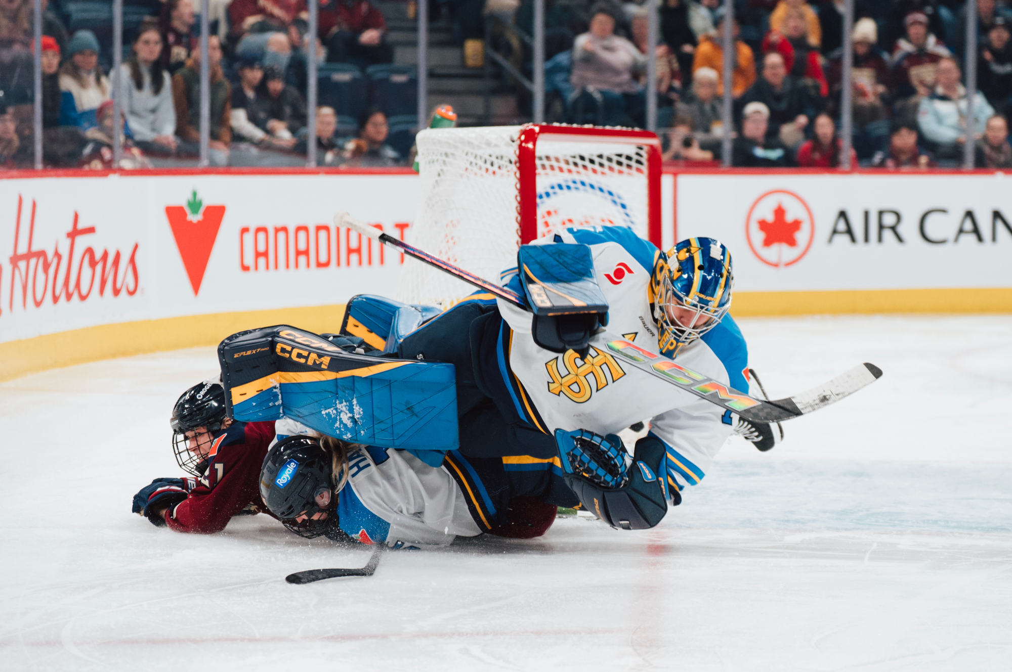 A Montréal and Toronto player are underneath a nearly horizontal Kirk as she attempts to make a save just outside her crease. The Montréal player is in a maroon home uniform, while Kirk is in a white away one.