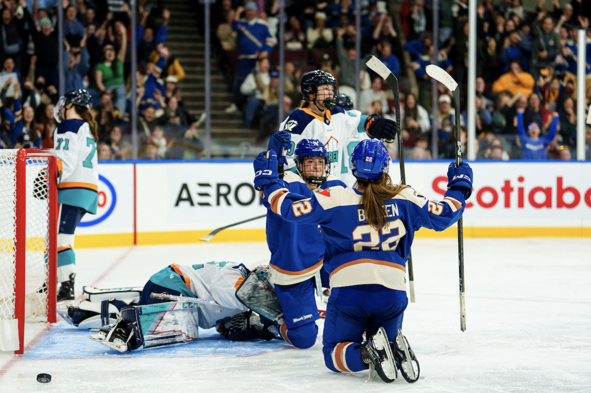 Boreen celebrates a goal with one of her teammates. Both are on their knees just outside the New York crease. The Vancouver players are in blue home uniforms, while the New York players in the background are in white away uniforms.