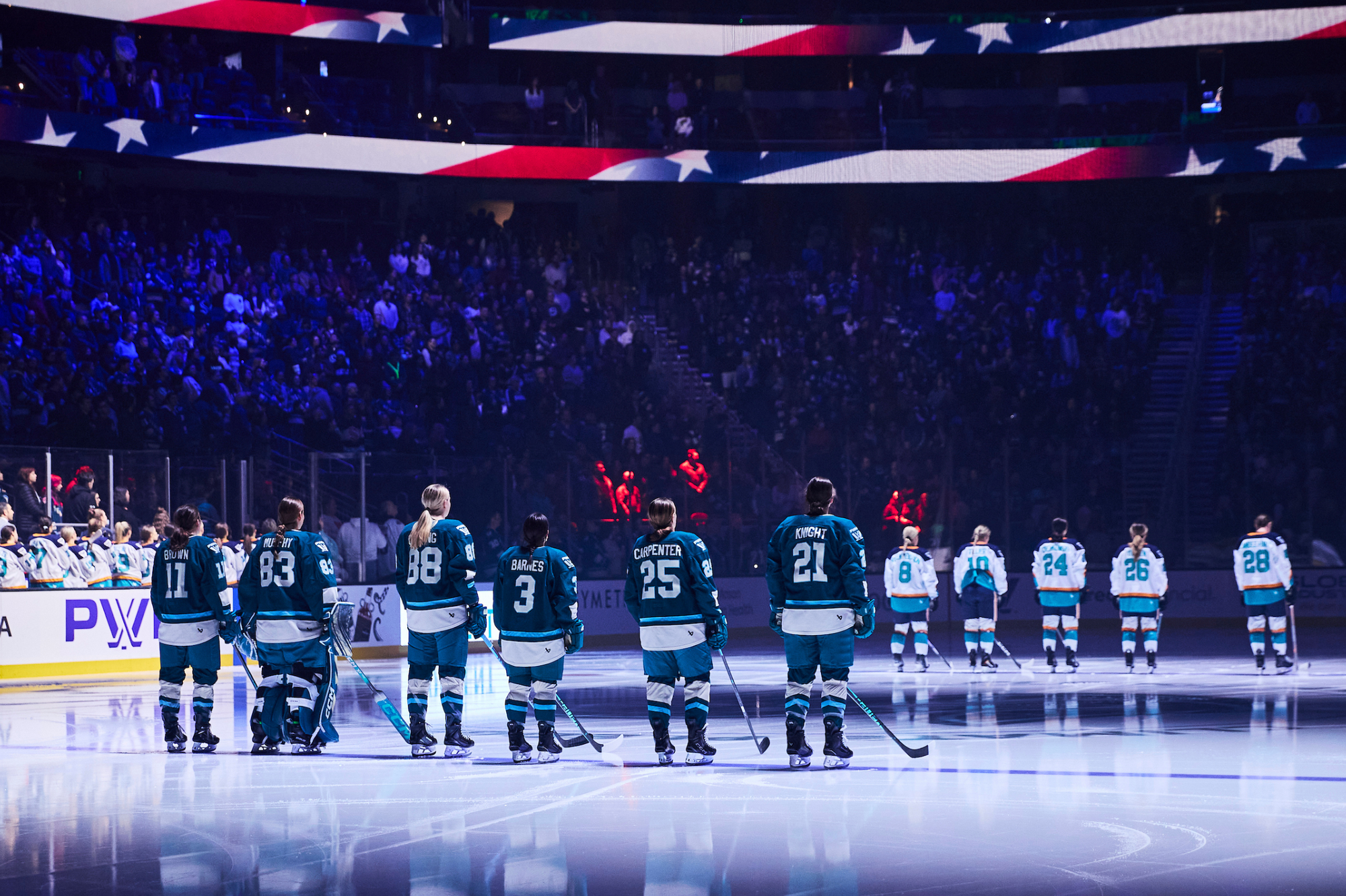 Six Seattle players are lined up on the blue line in the foreground, while five New York ones are visible in the background. All have their backs to the camera. The Seattle players are in teal home uniforms, while the New York players are in white away ones.