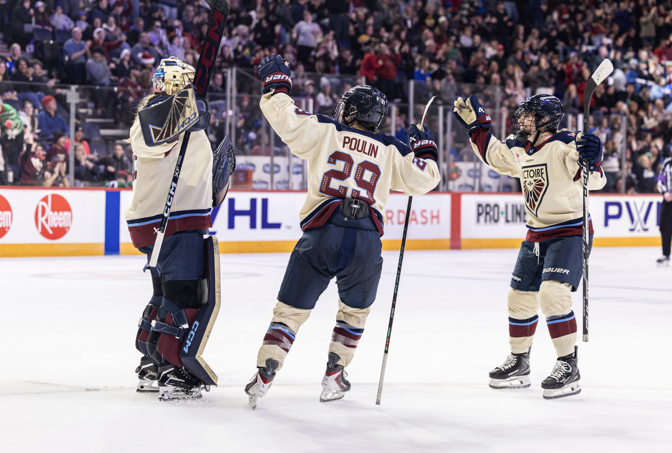 Desbiens, Poulin, and another teammate all raise their arms in celebration and come together for a group hug. They are wearing cream away uniforms.