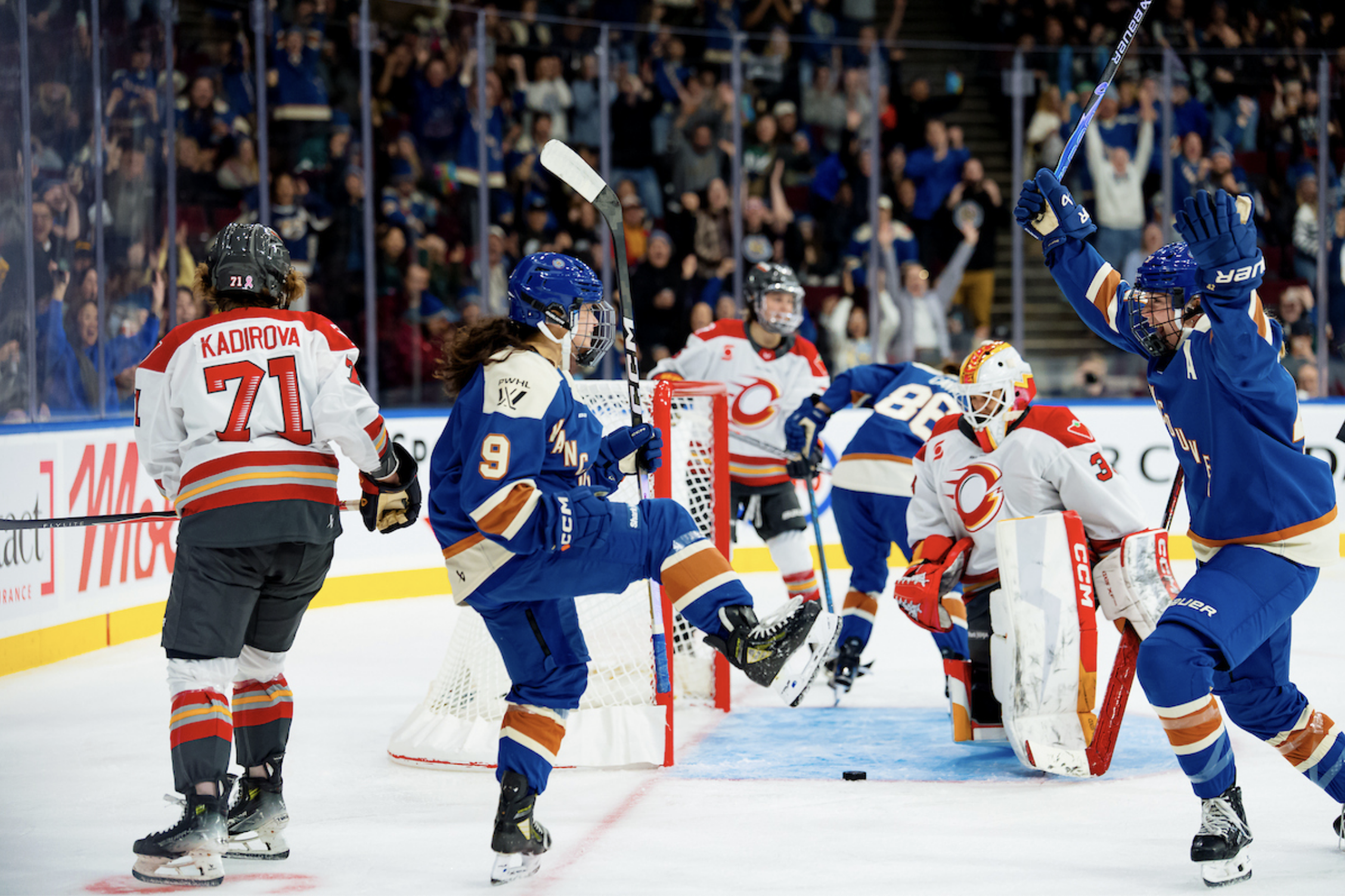 Chan raises her leg and pumps her fist in celebrate as one of her teammates skates toward her, smiling and arms raised. They are wearing blue home uniforms.