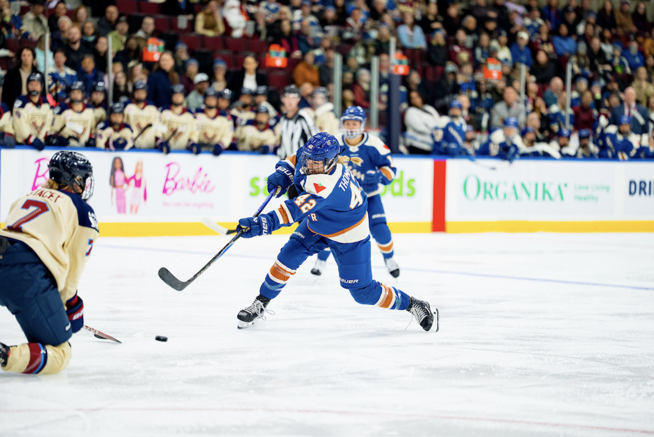 Thompson follows through with her stick as she fires a shot on goal as Montréal's Laura Stacey drops to a knee to block it. Thompson is wearing a blue home uniform, while Stacey is in a cream away one.