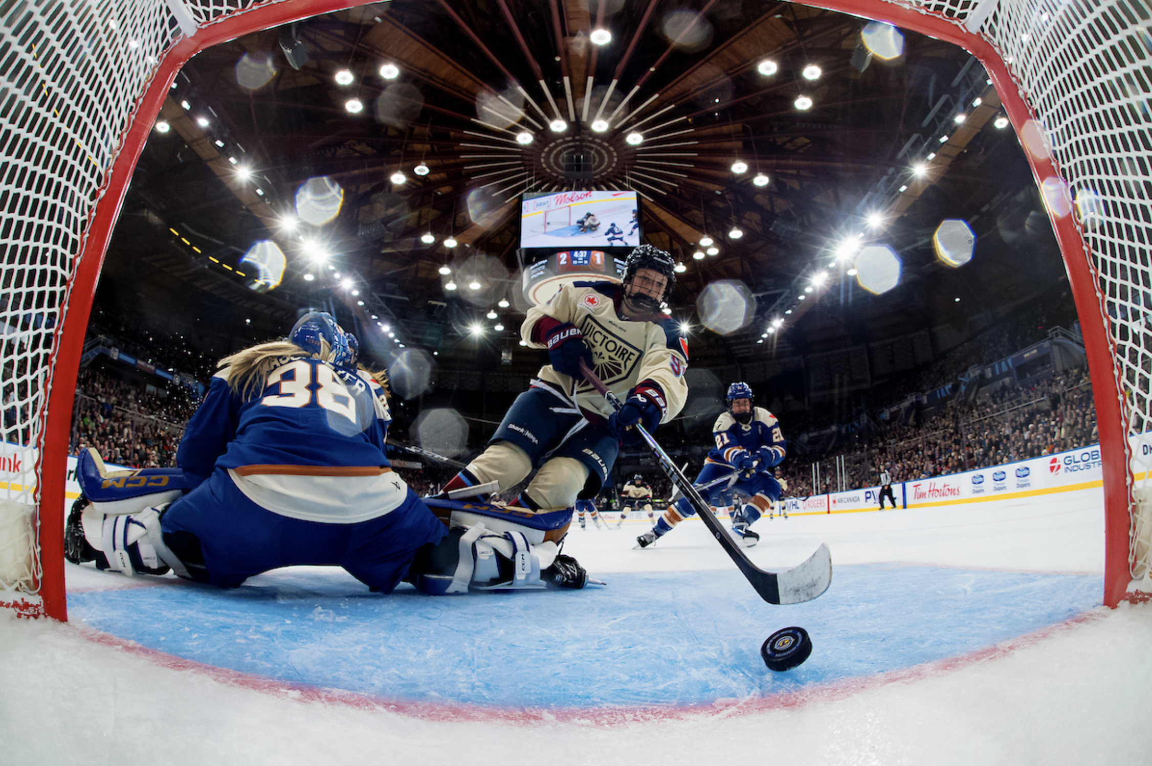 A shot from inside the net, as a Montréal player pushes the puck past Emerance Maschmeyer and over the goal line. The Montréal player is wearing a cream away uniform, while Maschmeyer is in a blue home one.