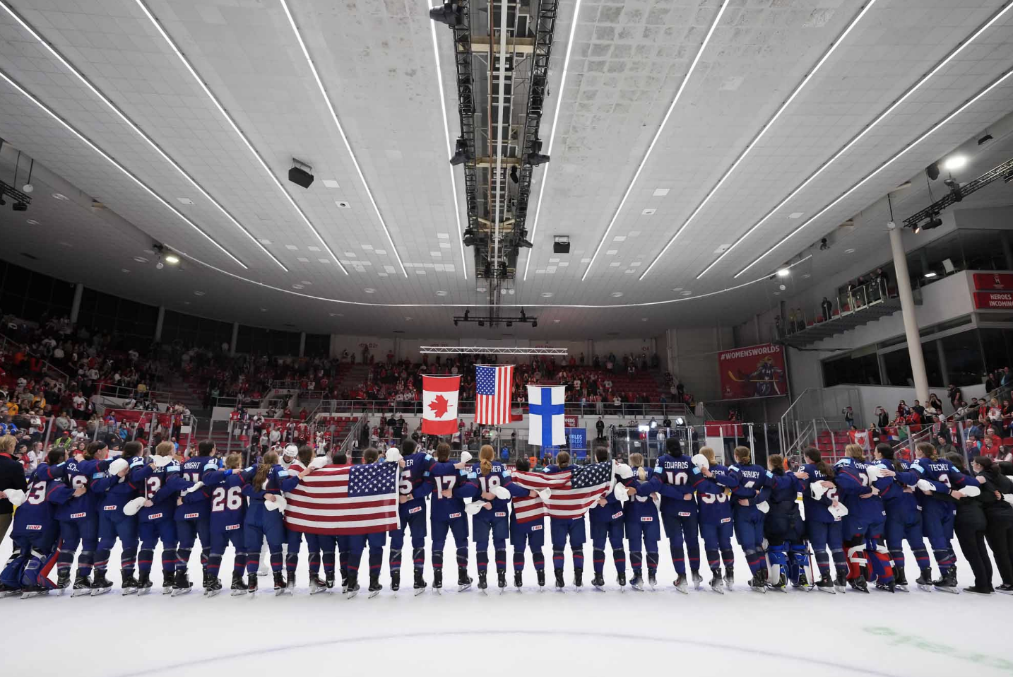 Team USA stands along the blueline, arm-in-arm, as the USA flag gets raised and the anthem plays to end the tournament. They are all wearing blue home uniforms. There are two American flags draped across some players backs in the middle of the line.