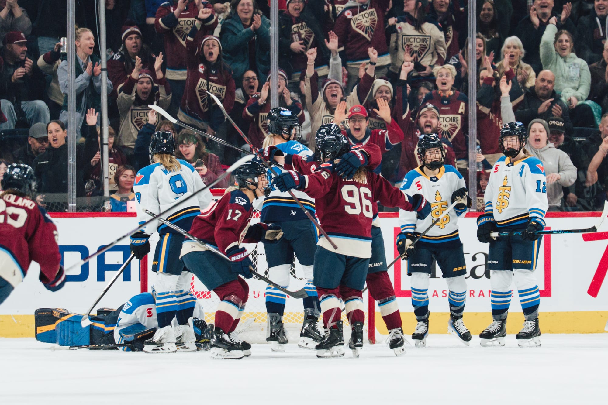 Victoire players come together for a group hug after scoring on the Sceptres. Part of the roaring crowd is captured in the background. The Victoire players are all in maroon home uniforms, while some dejected Sceptres players are standing in the background in white away ones..