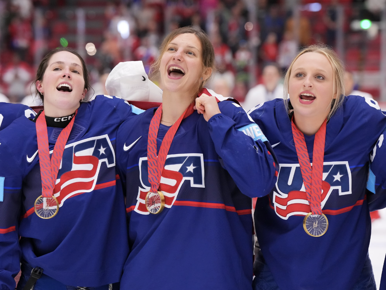 From left to right: Murphy, Eden, and Simms all smile wide while standing arm and arm, presumably singing along as the American anthem plays. They are wearing their gold medals around their necks and blue home uniforms.