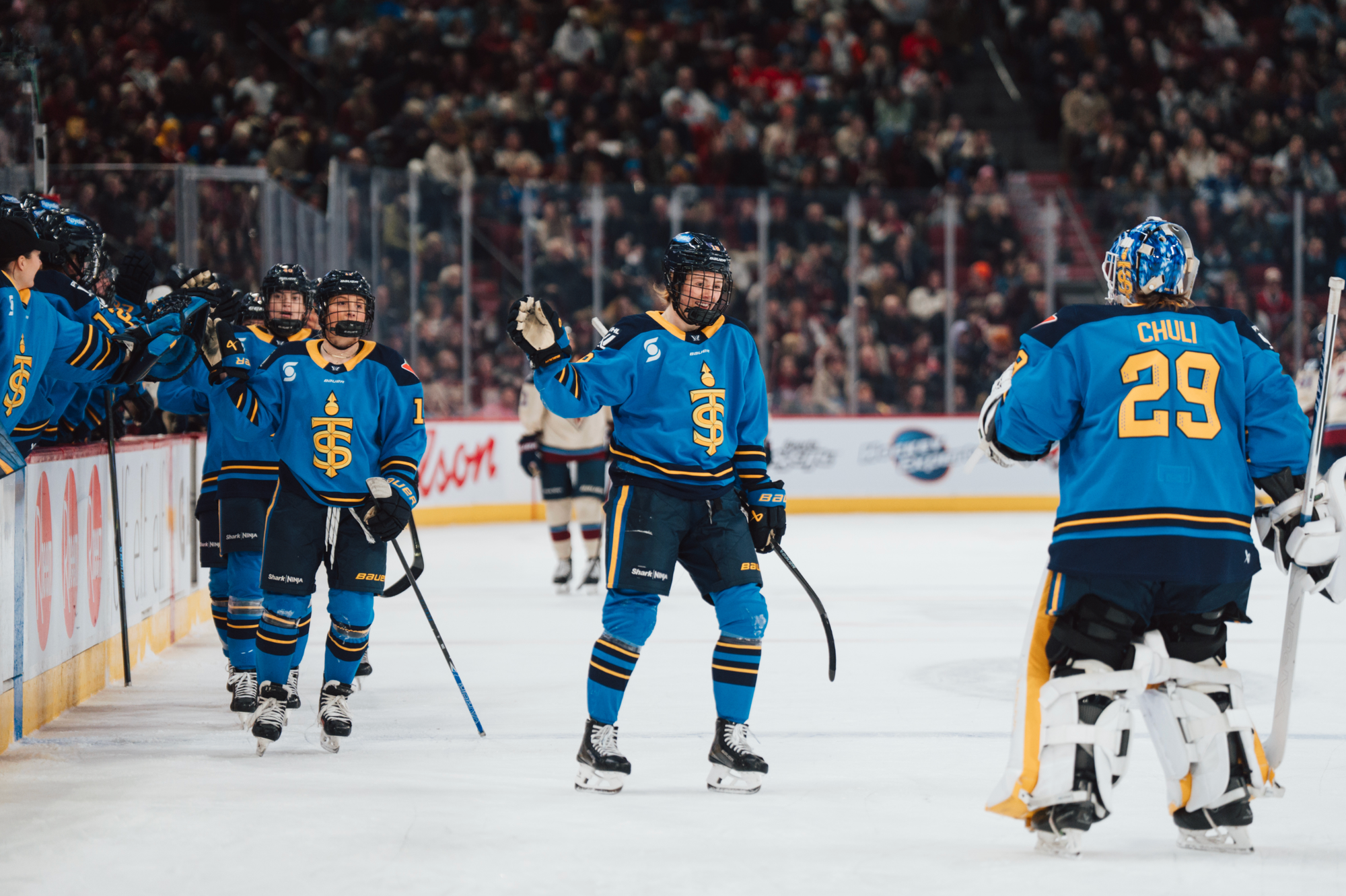 Compher and her teammates skate toward Chuli, who is by the blue line, after going down the bench handshake line. They are all wearing blue home uniforms despite being the away team in this game.