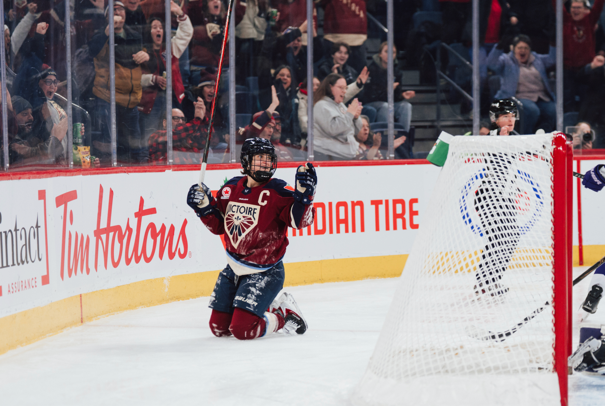 Poulin celebrates her goal against the Frost from her knees. She is behind the net with her stick raised, wearing a maroon home uniform.
