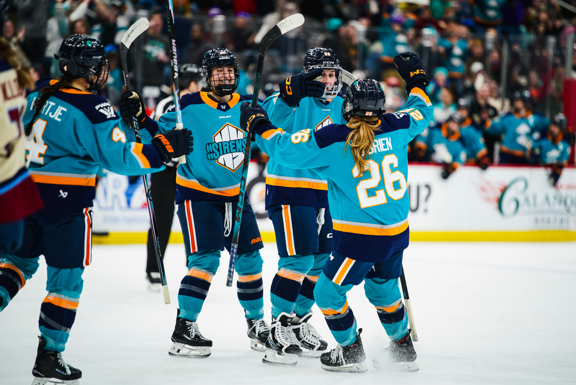 Four Sirens players come together to celebrate a goal with a group hug. They all have their arms raised. They are wearing teal home uniforms.