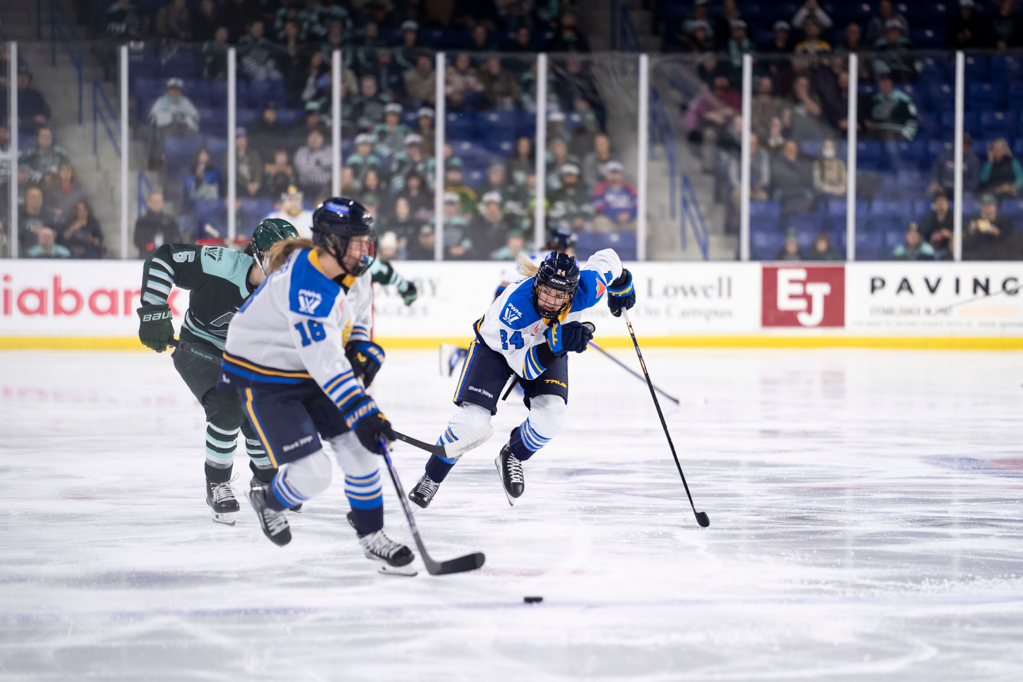 Compher, slightly out of focus, skates with the puck, as Spooner skates hard to catch up with her. A Fleet player is also hustling to defend the player. The Toronto skaters are in white away uniforms, while the Boston player is in a green home one.