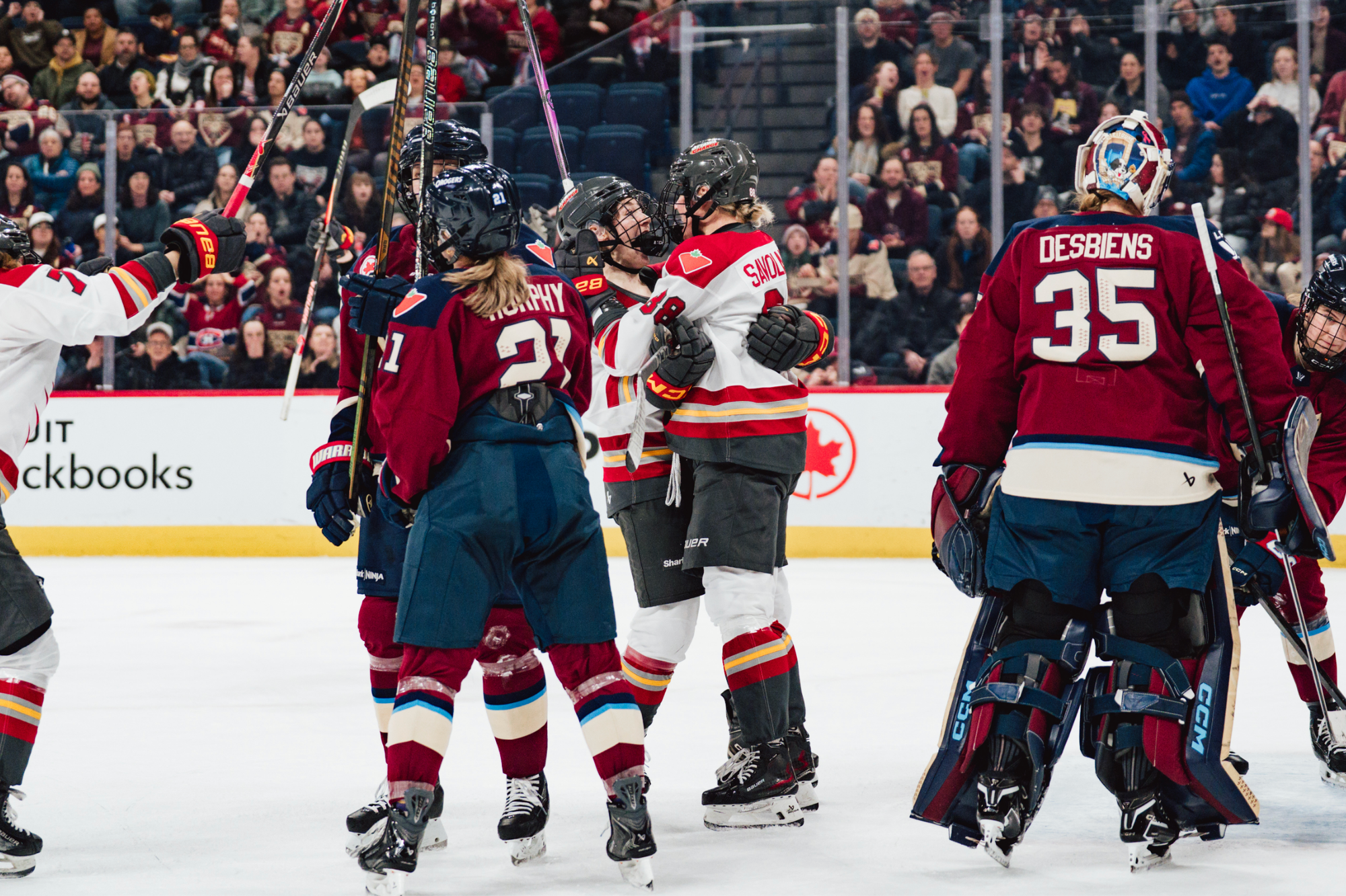 Savolainen hugs her teammate after scoring as others skate in to join from the outskirts of the frame. Several Montréal players dot the photo, wearing maroon home uniforms. The Charge players are all wearing white away ones.