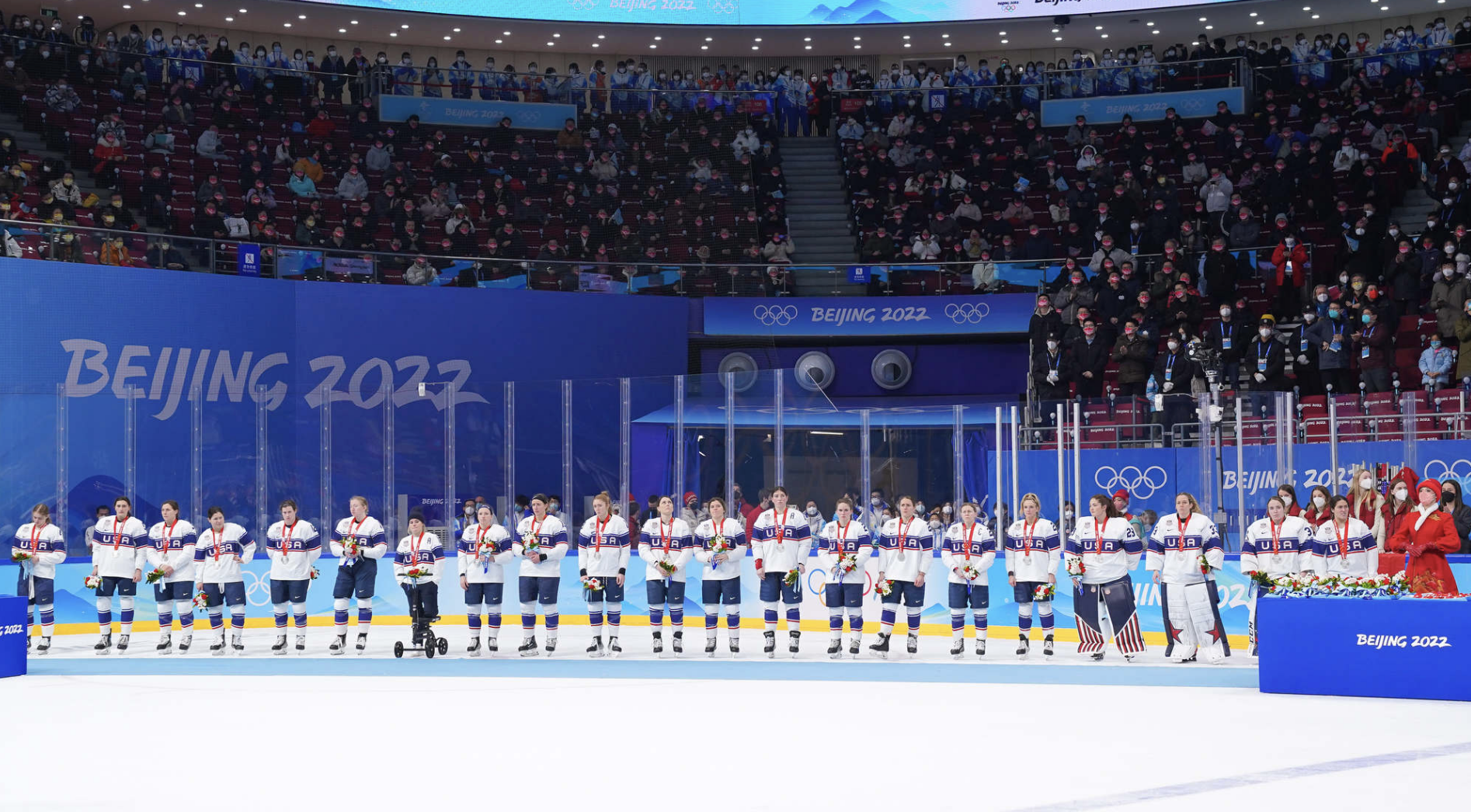 Team USA stands in a line, looking dejected with silver medals around their necks and flowers in their hands. They are wearing white away uniforms.